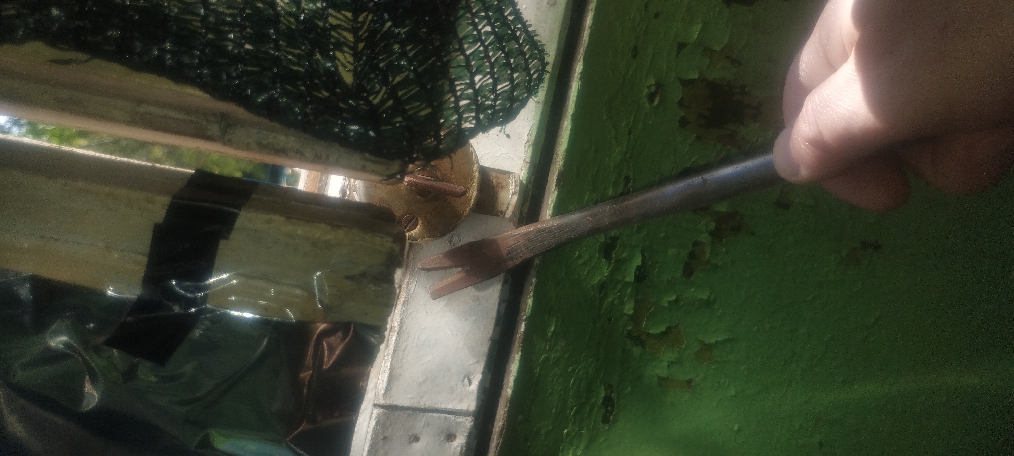 In Zaporizhzhia, Ukraine, a close-up shot captures a hand holding a metal tool, possibly a pry bar, against a green surface with peeling paint. The tool's tip, a U-shaped fork, is positioned near a white-painted wooden frame. Behind the frame, a dark plastic sheet is taped with black tape to a wooden support. A piece of green netting is also visible. The lighting suggests an outdoor environment, with sunlight casting shadows.