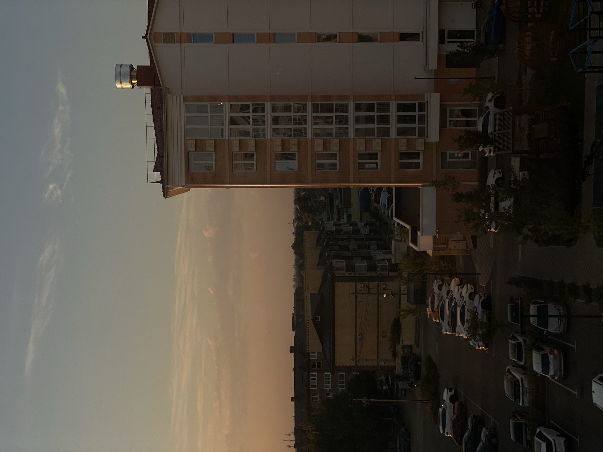 The image depicts a residential area in Vishnyaki, Russia, during what appears to be late afternoon or early evening, indicated by the warm, low-angled sunlight casting long shadows and the soft glow in the sky. In the foreground, a multi-story apartment building with orange brick and white siding dominates the right side of the frame. Its facade features numerous windows, many of which are large and reflective. A metallic cylindrical structure, possibly a vent or chimney, is visible on the roof.
To the left of the apartment building, a parking lot is filled with numerous cars, predominantly white, silver, and dark-colored sedans and SUVs. The cars are parked in marked spaces. Beyond the parking lot, several other apartment buildings of varying heights and architectural styles are visible, some with dark-tiled roofs and numerous windows. Trees and foliage are interspersed between the buildings and along the edges of the parking lot, with some trees appearing fuller and greener than others, suggesting varied species or levels of shade.
The background shows a hazy skyline with more buildings and trees in the distance, indicating a relatively built-up area. The sky above is a gradient of pale blue and warm orange-yellow hues, with scattered, wispy clouds. The overall scene is a typical urban residential landscape characterized by apartment blocks, parked vehicles, and vegetation under the gentle light of the setting sun. No people are visible in the image.