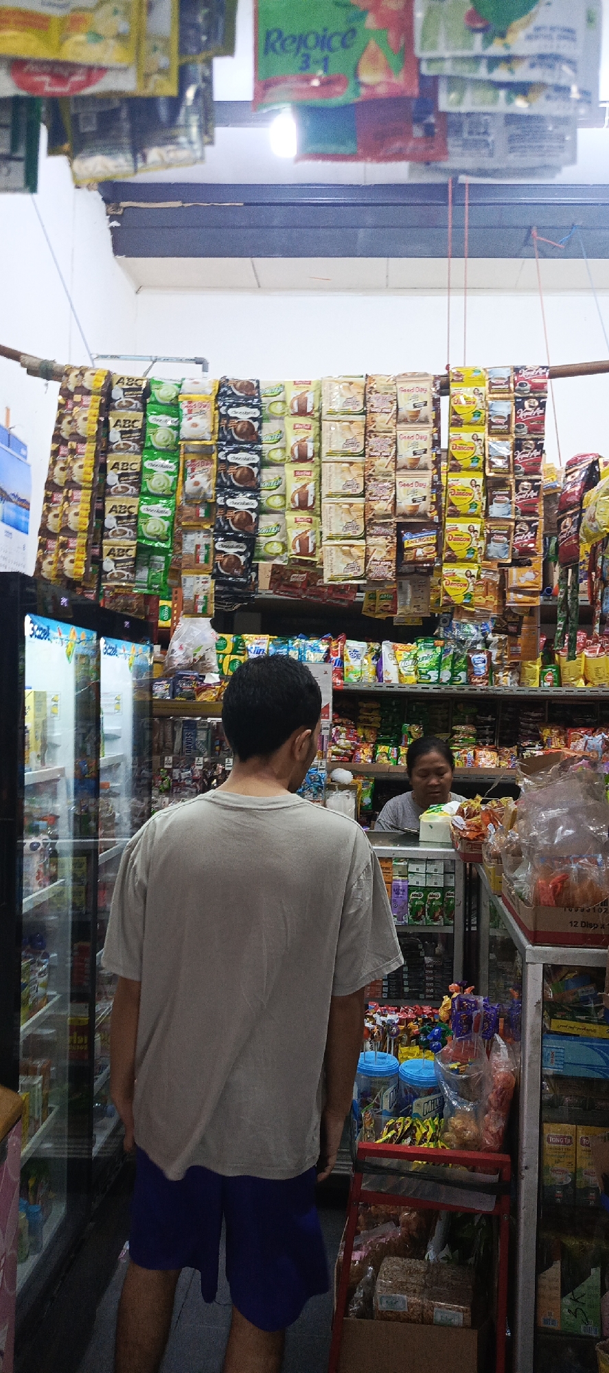 The image depicts the interior of a small convenience store, likely a warung, in Sukaresmi, Indonesia. The scene is dominated by shelves and racks filled with packaged goods, predominantly snacks and instant food items. In the foreground, a man wearing a grey t-shirt and blue shorts stands with his back to the camera, facing the interior of the store. To his right, a woman with dark hair, wearing a grey t-shirt, is visible behind a counter, interacting with the man or attending to the store. The store environment appears to be densely stocked, with items hanging from the ceiling and displayed on various shelves. A glass-doored refrigerator is visible on the left side of the image. The overall impression is a typical scene in a local Indonesian retail setting.