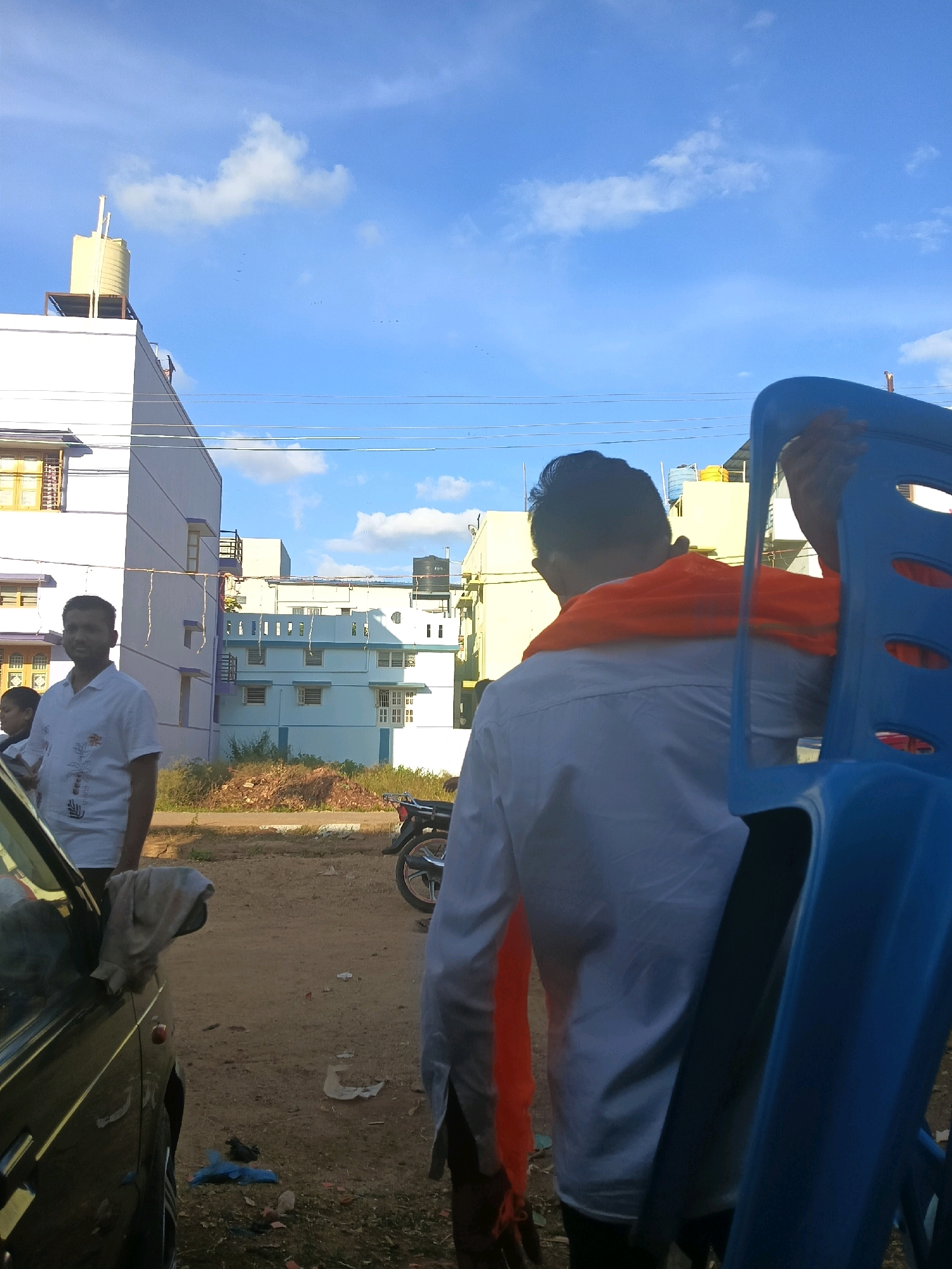 The image captures an outdoor scene in Jagaluru, India, under a bright blue sky with scattered clouds. In the foreground, a person seen from the back wears a white shirt and an orange cloth draped over their shoulders. They are holding a blue plastic chair, obscuring part of their body. To the left, another person in a white short-sleeved shirt stands facing the camera, with a portion of a black vehicle visible in the lower left corner. Behind them, a multi-story building with white and light blue facades and visible windows stands under construction or renovation, indicated by exposed plumbing and scaffolding. Further in the background, other buildings and a water tank on a rooftop can be seen. The ground is a dirt path with some debris, including scattered papers and plastic bags. A motorcycle is parked on the right side of the path. The overall environment appears to be a semi-urban or developing area.