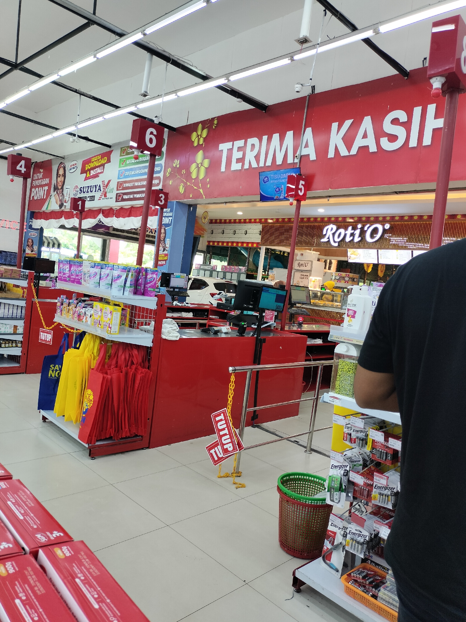 The image depicts the interior of a convenience store, likely a minimarket, in Langsa, Indonesia. The store features bright fluorescent lighting and red accents on its fixtures and signage. A prominent red sign across the top reads "TERIMA KASIH" (Thank You). Several numbered signs, indicating checkout lanes, are visible: "4", "6", "5", and "3". Lane 5 has a small screen displaying a product advertisement.
In the foreground, to the left, are rows of red-boxed products. In the center, a checkout counter area is visible with red displays and shelves stocked with various items, including reusable shopping bags in red and yellow. A "TUTUP" (Closed) sign hangs from a yellow chain.
Behind the checkout area, shelves are stocked with household goods like laundry detergent and toilet paper. Further back, a food counter with the "Roti'O" logo is visible, suggesting a bakery section within the store. A white car is parked just outside the store's entrance in the background, which appears to be a wide, open space with trees.
To the right, a person wearing a black t-shirt is partially visible, standing near a display of batteries. A brown woven basket sits on the floor next to a shelf displaying batteries and other small items. The floor is tiled with light-colored square tiles. The overall impression is a well-lit, organized retail environment.