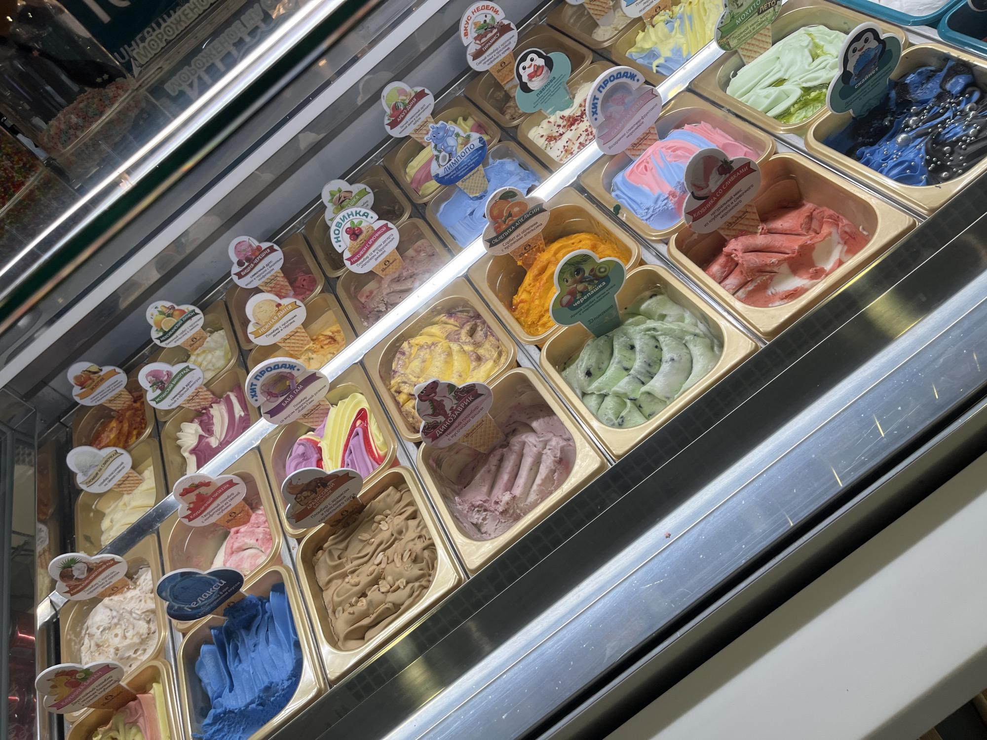 The image shows a display case filled with various flavors of gelato in individual containers. The display case is illuminated from above by fluorescent lights. Each container of gelato has a small label with Russian text and a picture, indicating the flavor. The flavors appear to be a mix of fruit, cream, and novelty options. Some of the labels are partially obscured by the gelato or other labels. The gelato itself is presented in scoops and swirls. The display case has a metallic frame and a glass front. In the background, out of focus, are more shelves with colorful toppings, likely for ice cream. The overall impression is of an ice cream or gelato shop. The image was taken in Sochi, Russia.