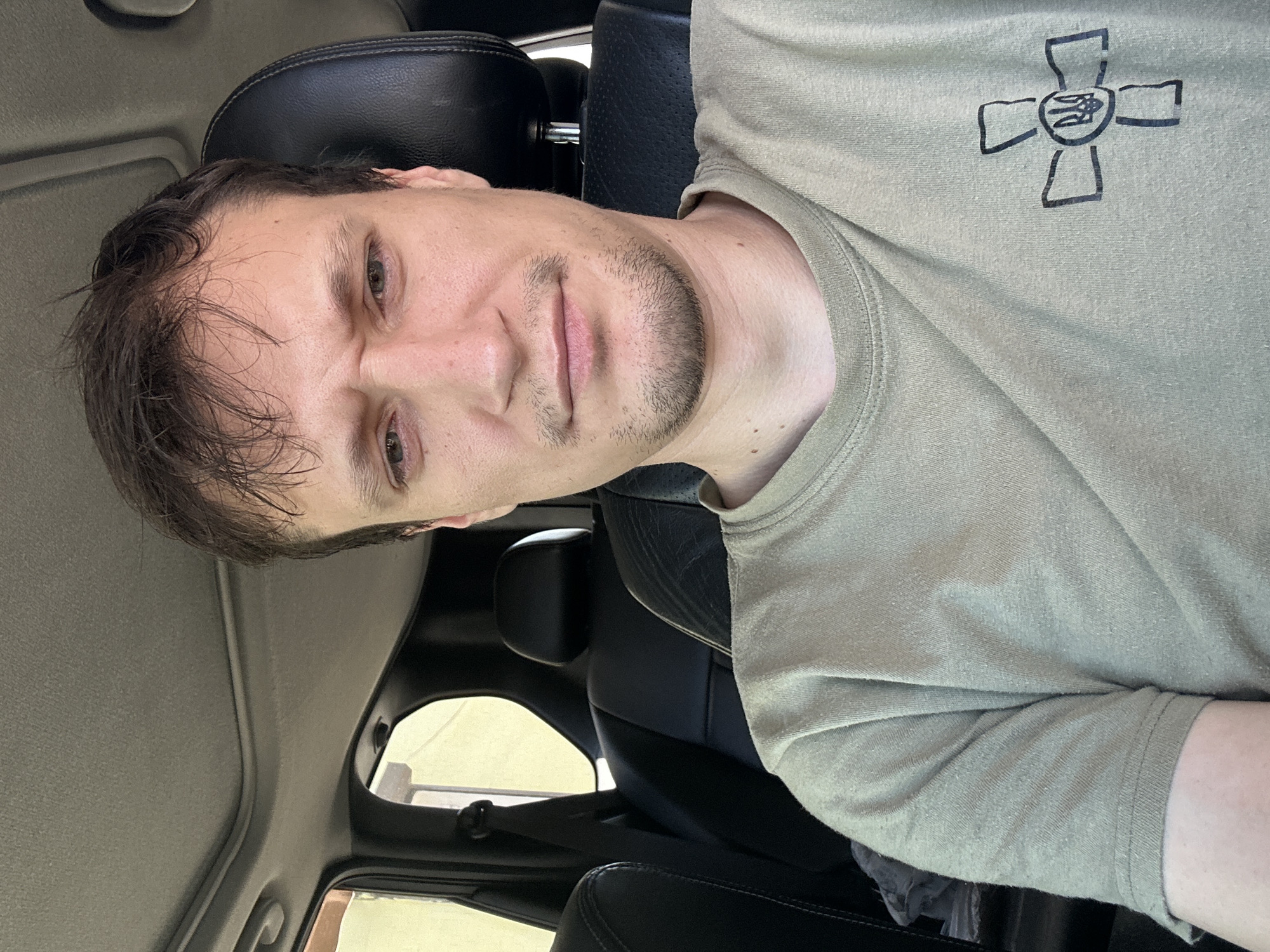 A man with short, dark hair and a slight stubble is sitting in the driver's seat of a car, looking at the camera. He is wearing a tan t-shirt with a black symbol of a cross with a trident in the center printed on the left side of the chest. The interior of the car is visible behind him, including the black leather seats and a sunroof. The lighting suggests it is daytime, and the background outside the car windows appears to be a bright, hazy yellow, possibly indicating an outdoor location. The image was captured in Shatava, Ukraine.
