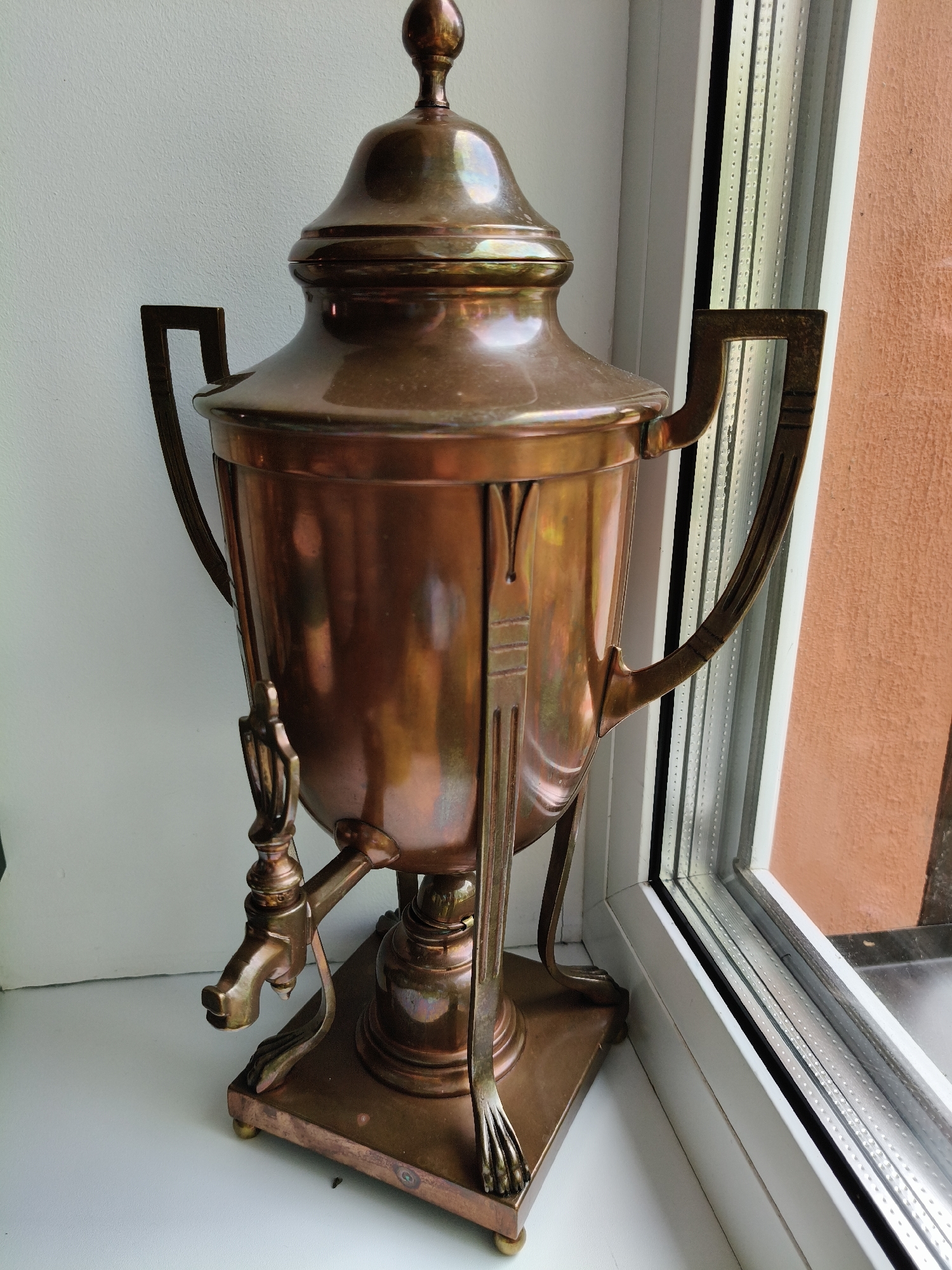 A vintage copper samovar is positioned on a white windowsill, adjacent to a window. The samovar, with its ornate handles and spigot, exhibits a warm, metallic sheen due to its material and the ambient lighting. The window, framed in white, reveals a textured, orange-toned exterior wall outside and a view of a grey, possibly overcast sky through the slatted blinds. The overall environment suggests an indoor setting, likely a home in Bigauņciems, Latvia, given the presence of the window and the exterior wall. No people are visible in the image.
