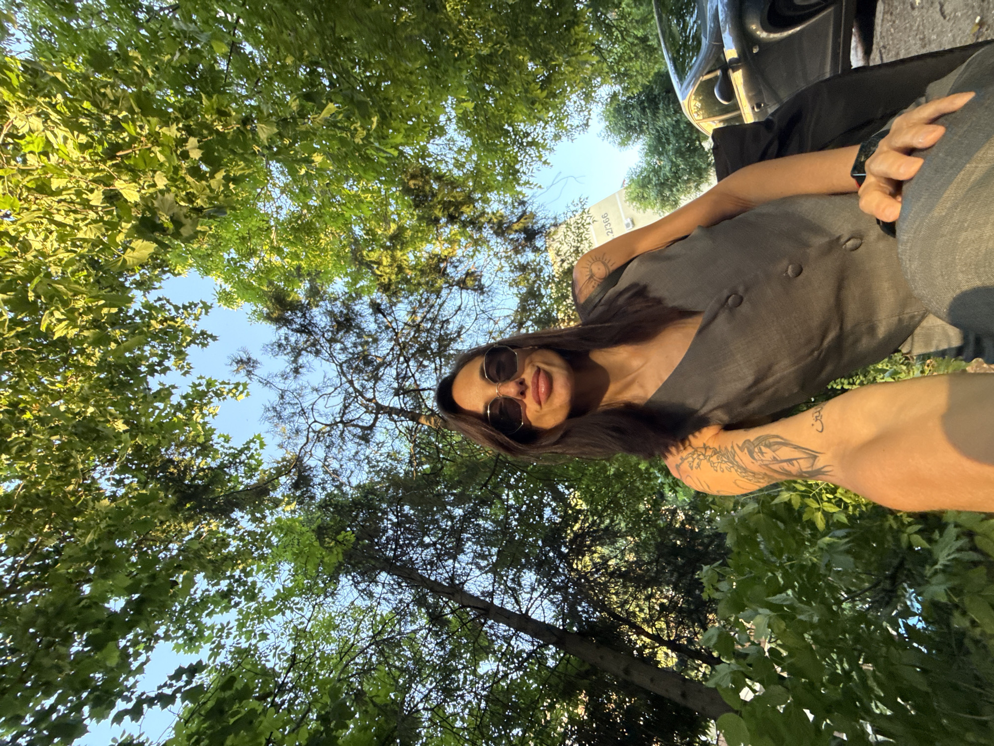 A low-angle, selfie-style photograph shows a woman outdoors with lush green trees filling most of the frame, suggesting she is standing in or near a wooded area or park. The woman has dark hair, sunglasses, and is wearing a grey sleeveless top. The presence of a partial image of a dark-colored car in the upper right suggests an urban environment. A building with an address "2/366" is visible. The exact location in Pleven, Bulgaria, cannot be definitively determined from the image but based on the style of architecture visible in the address “2/366” it is very possible this photo was taken near the Pleven Panorama.