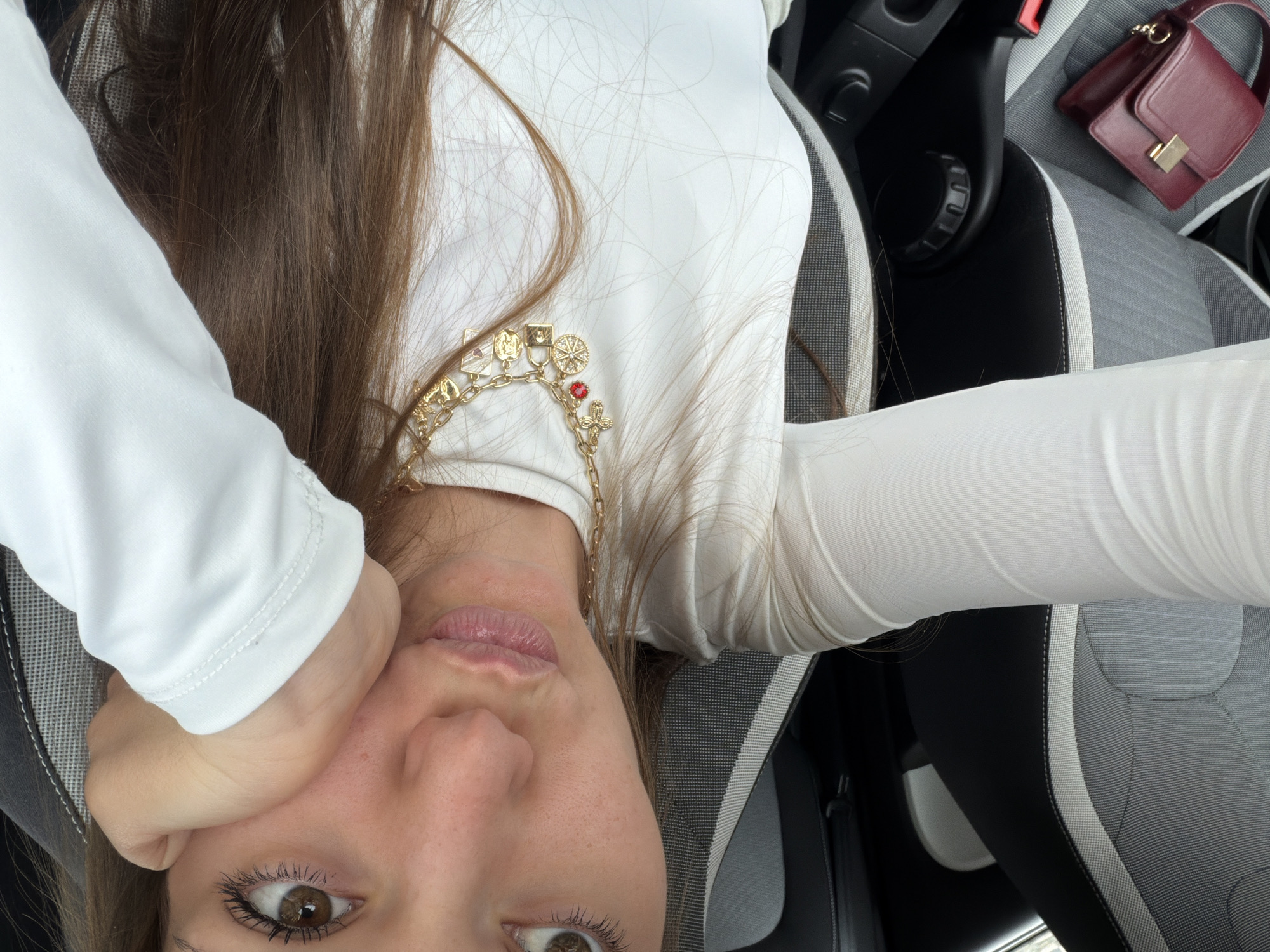 A selfie photograph shows a woman inside a car. She is wearing a white, long-sleeved shirt and a gold charm necklace. Her hair is brown. Her face is partially obscured by her hand, and she is looking towards the camera. A small, burgundy purse is visible on the car seat. The car interior is gray and black. The location cannot be determined from this image.