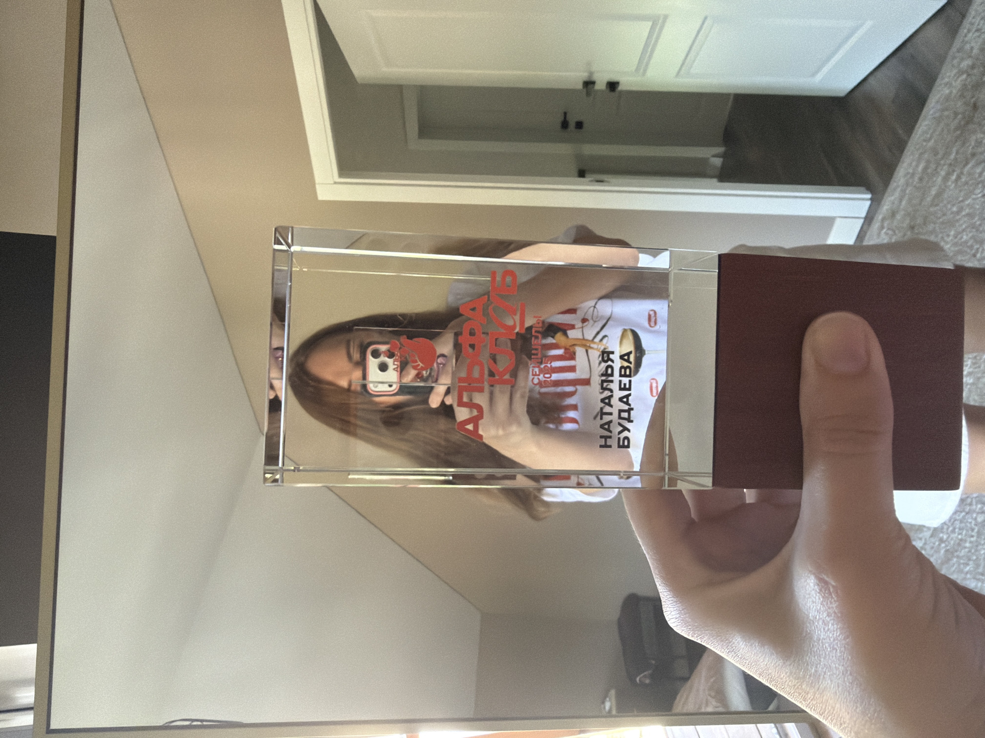 A person's hand holds a glass trophy with a red base. The trophy is in focus, and a reflection in the trophy shows a woman holding a phone, taking a picture. She is wearing a white t-shirt with red lettering that reads "АЛЬФА КЛУБ" (ALPHA CLUB) and "НАТАЛЬЯ БУДАЕВА" (NATALIA BUDAEVA). The background is a blurred interior of a room with a mirror, a door, and a light-colored wall. The scene appears to be indoors, possibly in a residential setting. There is no visual information to suggest the location is specifically Malinovka, Russia.