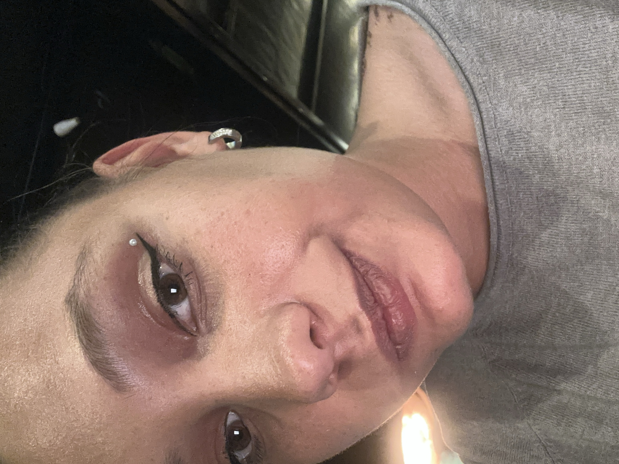 A close-up, rotated headshot of a fair-skinned person. The person has an eyebrow piercing on their left eyebrow, an earring on their left ear, brown eyes, and full lips. They are wearing a grey top. The background appears to be dark. There is no indication of the location being Sofia, Bulgaria.