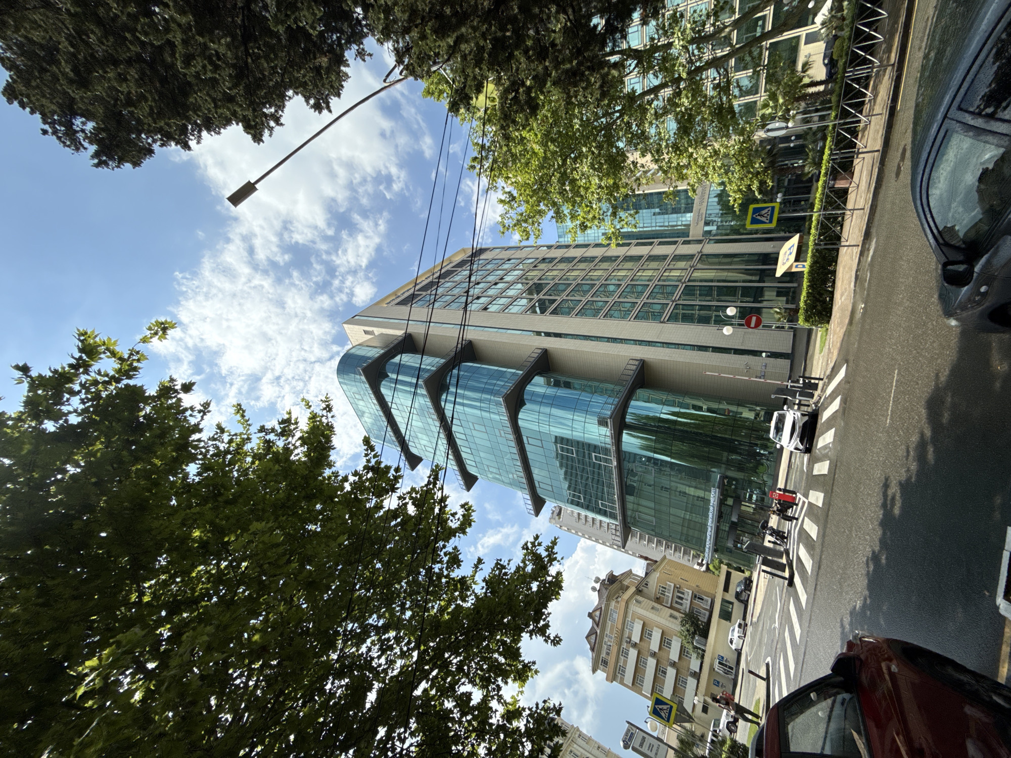 This is a street-level, medium-angle, full-shot photograph taken on a sunny day in Sochi, Russia, featuring a modern, multi-story glass-fronted building with a curved facade, possibly an office building or hotel, and an adjacent, older, multi-story building with beige siding and a pitched roof. The foreground shows a wet asphalt road with white crosswalk markings. A dark red car is partially visible in the lower left, and a dark gray car is in the lower right. Several other cars and two motorcycles are on the road and near the intersection. Pedestrians are visible on the sidewalk near the entrance to the glass building. Trees with green foliage frame the scene from the left and right, with the sky and clouds visible above. Streetlights and traffic signs are also present. The building's facade reflects the sky and surrounding trees. The inscription on the glass building appears to read "РОССИЙСКИЙ МЕЖДУНАРОДНЫЙ ОЛИМПИЙСКИЙ УНИВЕРСИТЕТ".