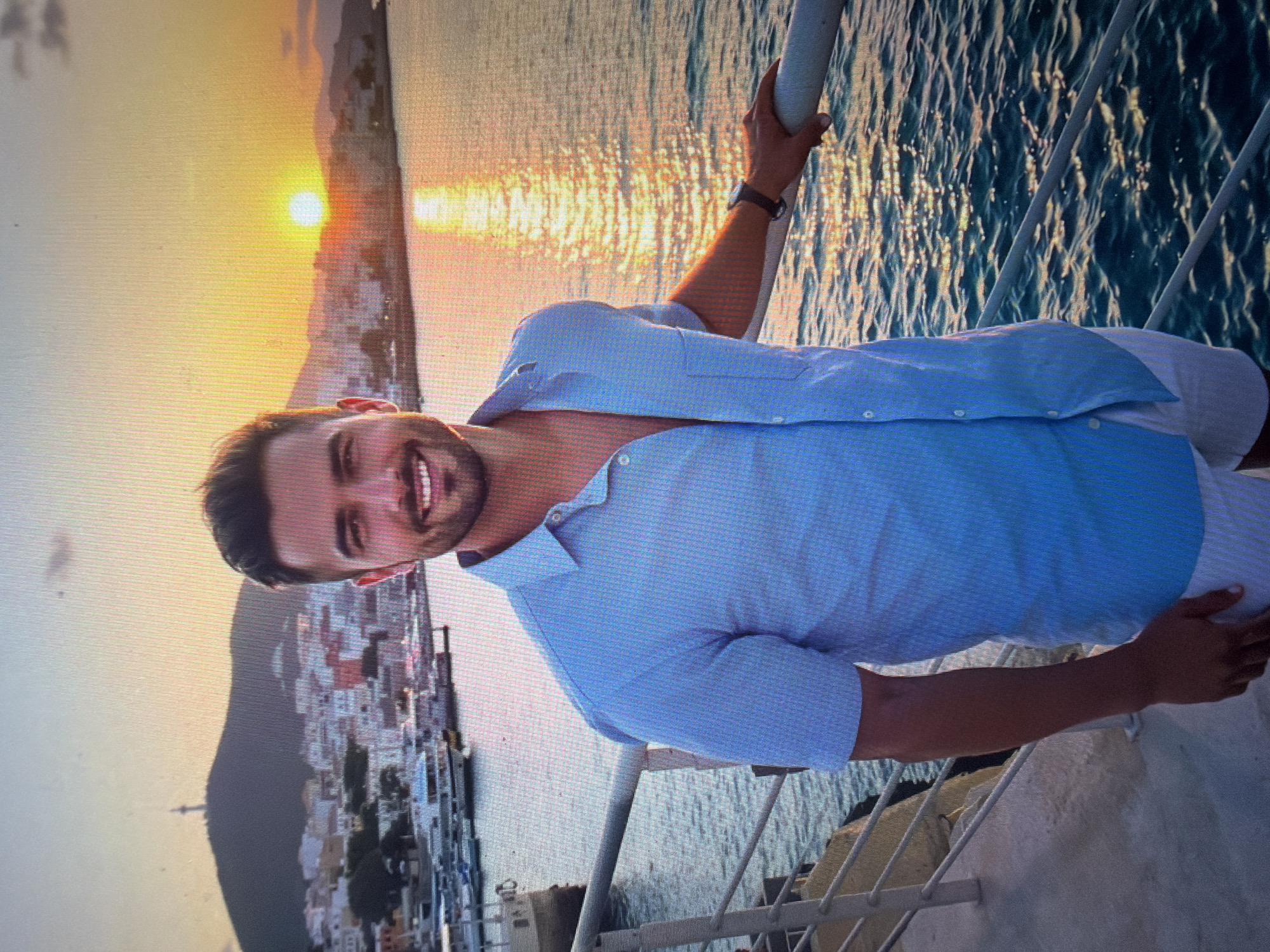 The image captures a smiling male individual, appearing to be in his 20s or 30s, positioned in the foreground. He has dark hair and is wearing a light blue, short-sleeved, collared shirt, partially unbuttoned, and white shorts. His right hand rests on a white metal railing that spans across the lower mid-section of the frame, and a dark-strapped watch is visible on his left wrist. The scene is set at sunset, with a bright orange sun visible on the horizon to the right, casting a strong golden reflection across the water. In the background, a bustling coastal city, identified as Shumen, Bulgaria, stretches along the waterfront, featuring numerous boats and yachts docked in a harbor. Behind the cityscape to the left, a prominent hill or mountain rises, topped with a distinctive, slender structure. The sky displays a gradient of warm colors, including orange, yellow, and light blue, indicative of dusk. The water in the foreground, beyond the railing, shows ripples reflecting the ambient light.
