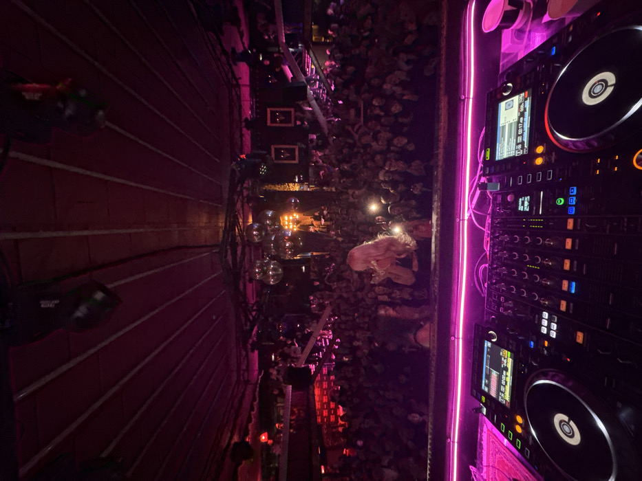 This image depicts the interior of the Tesla Club in Rostov-on-Don, Russia, during a live event. The perspective is from the elevated DJ booth, looking out over a densely packed crowd towards the main stage.
In the immediate foreground, a professional DJ setup is visible, featuring two Pioneer CDJ-3000 media players and a Pioneer DJM-900NXS2 mixer. The console surface is illuminated by a prominent magenta neon light strip.
The midground is dominated by a large audience, estimated to be several hundred individuals, filling the main floor and occupying elevated sections on the left and right. Many attendees are actively holding up smartphones, with multiple devices emitting bright white light from activated flashlights or screen illumination, directed towards the stage. A woman with long blonde hair is prominently positioned near the front of the crowd, her back to the camera, appearing engaged with the performance.
The background reveals the stage area, distinguished by several large, illuminated disco balls suspended from the ceiling. Bright, warm-toned stage lighting emanates from the center of the stage. The club's ceiling is vaulted, displaying visible structural beams, all bathed in a magenta light consistent with the overall lighting scheme. Elevated platforms or balconies are situated on both sides of the venue, also populated by audience members. Decorative elements include two dark-framed pictures hanging on a wall to the right, and a tall, dark, tree-like structure positioned to the left of the main stage.
The overall environment is characterized by low ambient light, primarily magenta and purple, punctuated by bright stage lights and the white light from numerous smartphone flashlights, creating a vibrant club atmosphere during a performance.