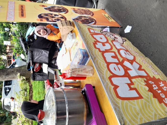 The scene is set up on a sidewalk food stall, with a large sign that reads "Mentai Ker" with "Serba 5k" beneath it, in Kecamatan Sidoarjo, Indonesia (latitude: -7.442, longitude: 112.705). Two people are present: a woman in a black hijab is standing behind the stall, and a woman wearing a hijab in orange and blue is sitting beside a motorcycle parked in front of the stall. The stall features a metal stand with a double-layered steamer pot, a small stove with a purple base, and a variety of spices and sauces. Other objects in the scene include a large black bag on the motorcycle, several take-out containers, plastic straws, and a large white sign behind the stall with the text "DIMSUM MENTAL Harga Mulai 15.000". The environment is casual and street-like. The stall is selling food items that are not visible in the image.