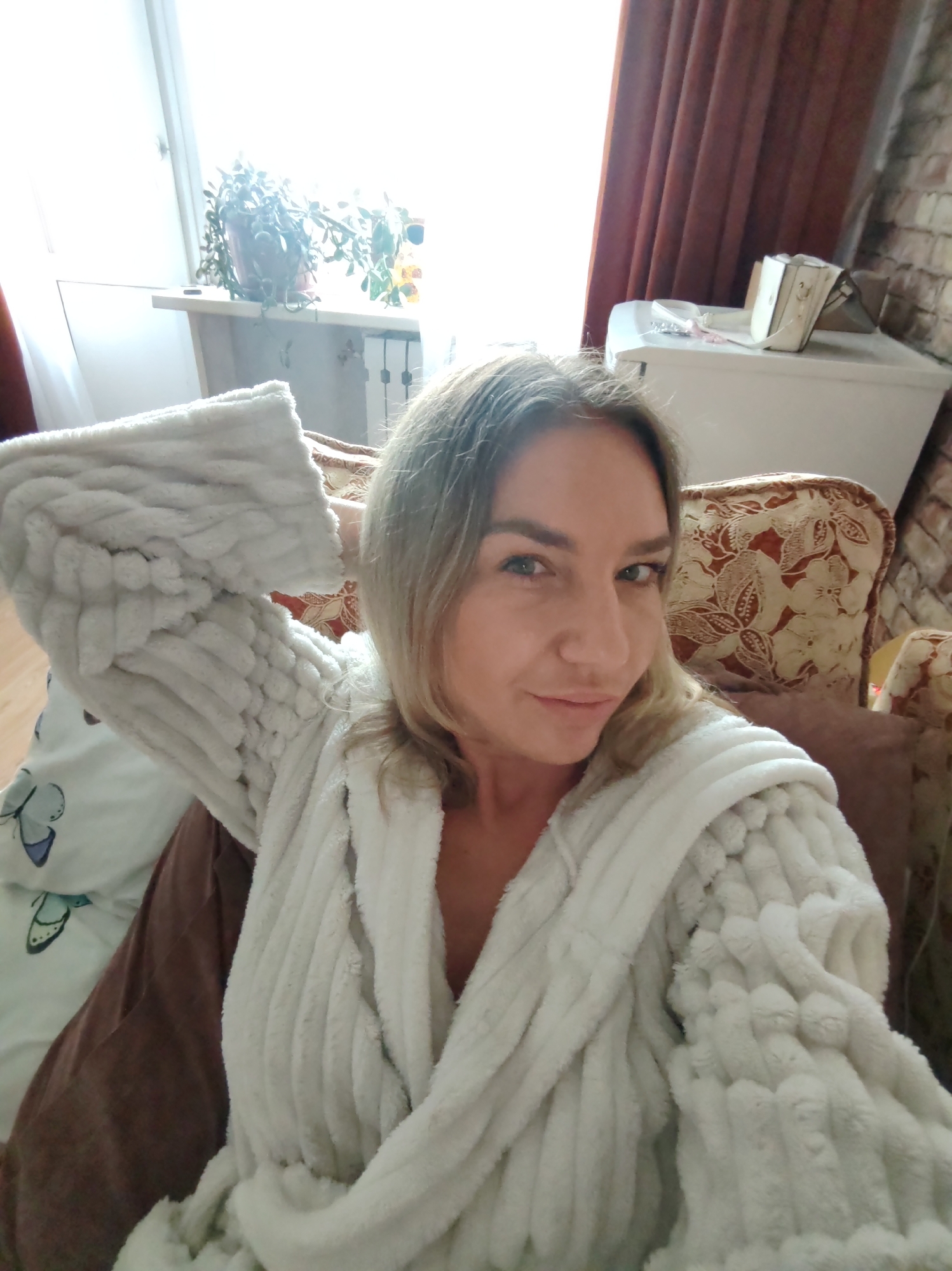 A woman in a white robe is the primary subject. The background includes a window with a plant, a patterned chair, and a white cabinet. The location cannot be determined as Petropavlovsk-Kamchatsky, Russia, based solely on this image.