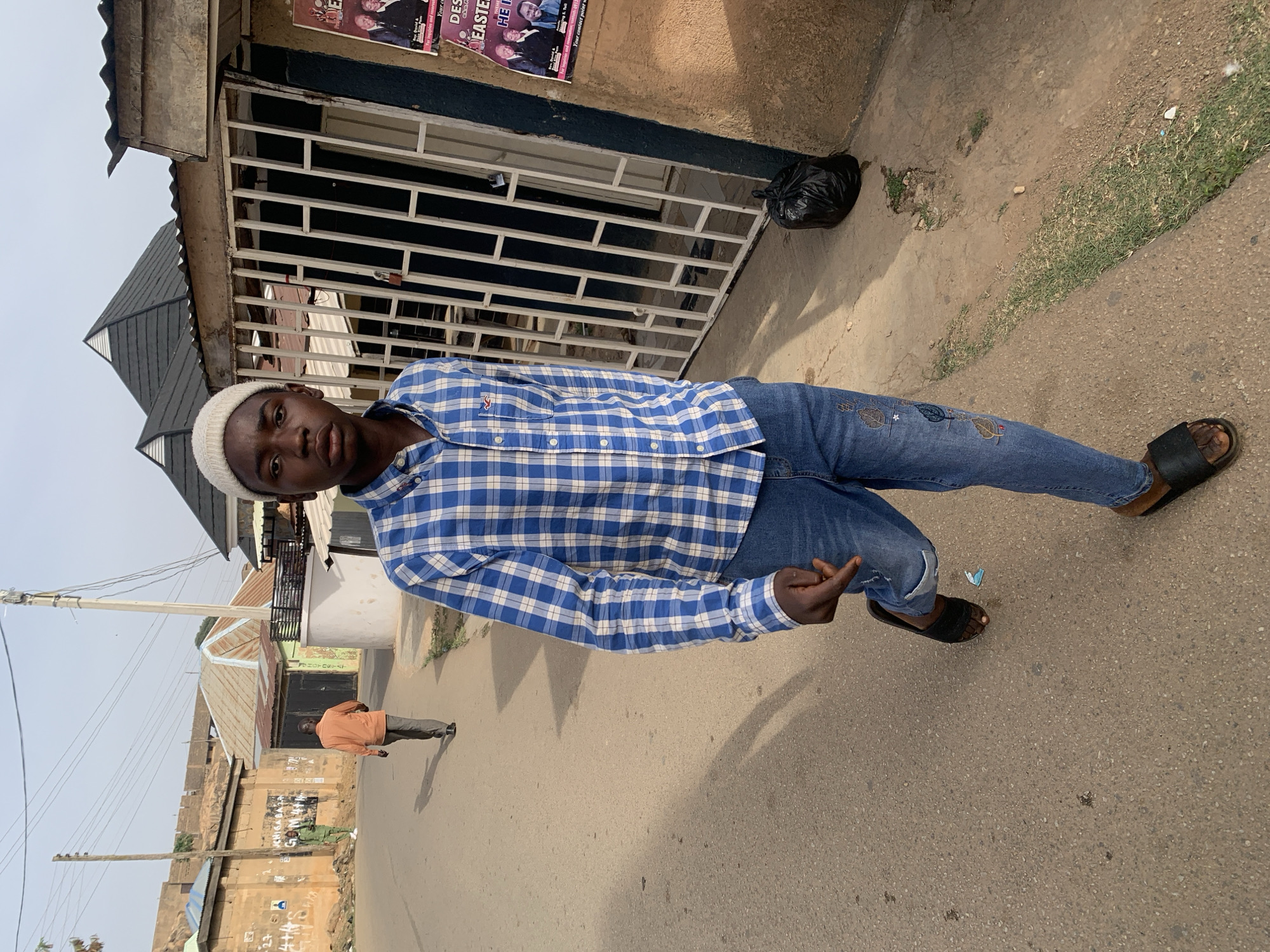 A young man stands on a street, angled towards the camera, wearing a white knit cap, a blue and white checkered shirt, and jeans with embroidered designs. He is also wearing dark-colored sandals, and his left hand is raised, showing the middle finger. Behind him, a building with a white metal gate covers the shopfront. Posters are visible on the building. A black plastic bag sits on the sidewalk near the building. Across the street, buildings are visible, and a man wearing an orange shirt and gray pants walks in the background. Utility poles and wires line the street. The street surface is asphalt. The location appears to be in a populated area, possibly the city of Jos, Nigeria.