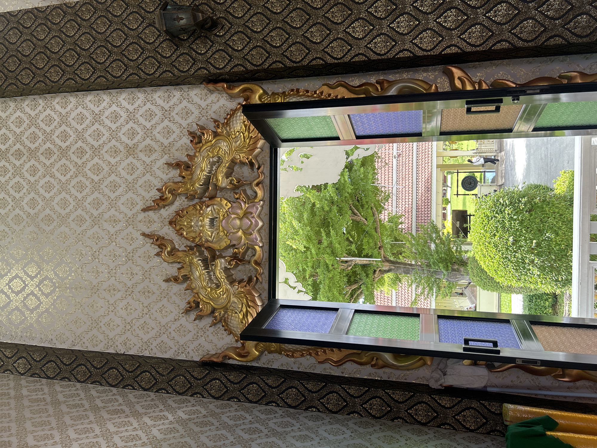 The image shows an ornate doorway with a golden dragon motif above it, opening onto a lush green garden. The doorway itself is framed by decorative wallpaper and dark patterned fabric panels. The open door reveals a view of a tiled roof building, a large tree, and manicured bushes. A person in white is visible in the distance on a paved path. The overall scene suggests an entrance to a temple or a similar place of cultural significance, possibly in Thai Ko Subdistrict, Thailand, given the style of the decoration and the architectural elements.