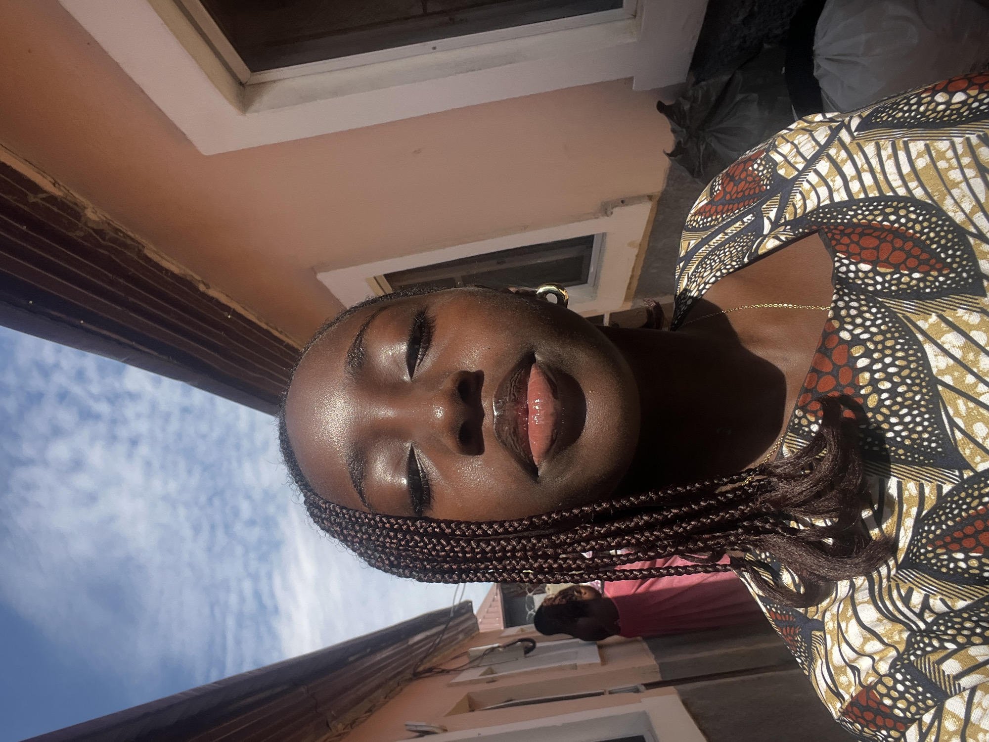 The image depicts a close-up selfie of a young woman in a narrow outdoor passage in what is described as the city of Abuja, Nigeria.
The primary subject is a woman with dark skin, her eyes closed, and her head slightly tilted back, basking in bright sunlight. Her lips are full and appear glossy. Her hair is styled in numerous thin, dark brown box braids, with a prominent section falling over her left shoulder (viewer's left). She wears a brightly patterned top or dress, featuring a mix of geometric and organic designs in earthy tones of gold, dark brown, black, white, and reddish-orange. A small gold hoop earring is visible on her right ear (viewer's right), and a thin gold chain necklace rests on her chest. Her skin has a visible sheen, indicative of either moisture or natural oils reflecting the strong sun.
The background shows two buildings with light peach-colored stucco walls, creating a confined corridor. On the left side, a section of a white window frame with dark glass is visible. Further back and to the left, another individual is partially visible, seen from behind, wearing a long pink or reddish-pink garment, possibly a dress or skirt. On the right side, another white window frame with dark glass is visible on the building wall. Above, the undersides of dark brown wooden eaves or soffits line the top of both buildings. The sky above is bright blue with scattered white, wispy clouds. In the lower right corner, several dark trash bags are visible on the ground against the building wall. The scene is illuminated by strong, direct sunlight, creating distinct highlights and shadows.