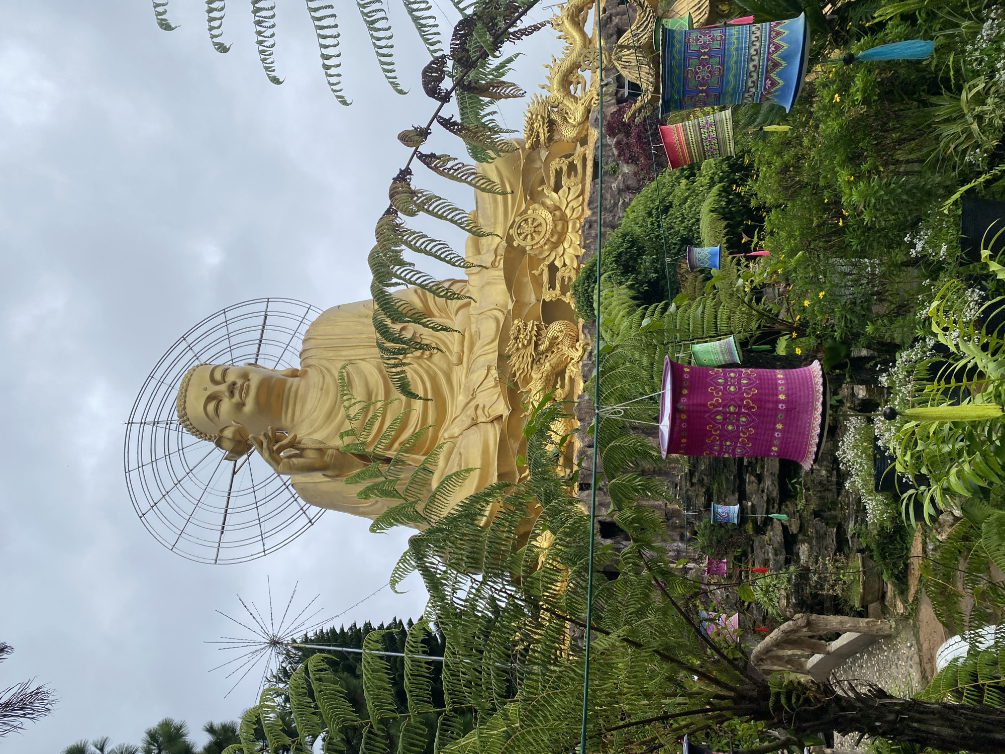 The image captures a large, golden Buddha statue seated outdoors in what appears to be a temple or garden setting in Lâm Viên - Đà Lạt, Vietnam. The Buddha is adorned with a circular halo or nimbus behind its head, and is depicted with its right hand raised, holding a flower. The statue is positioned on a rocky, tiered base, with ornate golden dragon sculptures flanking it. In the foreground, lush green ferns and other foliage create a natural frame for the scene. Several colorful, cylindrical lanterns, decorated with intricate patterns and tassels, hang from unseen supports, adding a festive touch to the environment. The sky is overcast, suggesting a cloudy day. The background reveals more greenery and architectural elements suggestive of a Buddhist sanctuary.