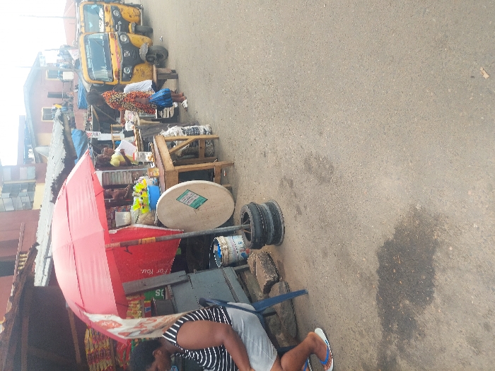 The image depicts a street scene in Agani, Nigeria, characterized by market activity along the left side of a paved road. In the foreground, a woman wearing a black and white striped top and light shorts is seated on a blue plastic chair, facing away from the camera. Her feet are adorned with white flip-flops. Adjacent to her are concrete blocks and two stacked black tires.
A red umbrella provides shade over a makeshift market stall, with a red banner partly visible beneath it, displaying "For You." A wooden table positioned to the right of the umbrella holds several plastic bowls containing yellow contents, alongside bottles and other small items. A large, circular, light-colored object with a green label leans against the table's leg. Another individual in a yellow top is partially visible behind this stall. The background on the left reveals various multi-story buildings with corrugated metal roofs, indicative of an urban setting.
The paved asphalt road, with a prominent dark stain in the lower-left foreground, extends toward the background. On the far right, three yellow motorized tricycles, commonly known as Keke Napep, are parked in a line. Several individuals are present near these tricycles; one person in a colorful patterned dress stands, while others are seated on small wooden stools. The overall lighting suggests daytime conditions.