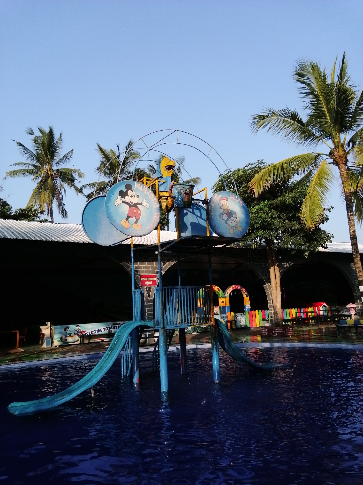 The image depicts a water park located in Kebonsari, Indonesia. In the foreground, a turquoise water slide curves into a dark blue swimming pool. The slide is supported by blue metal poles. Behind the slide, a multi-level structure, also painted blue, features circular panels adorned with cartoon characters: Mickey Mouse on the left and Donald Duck on the right, with the text "MUTIARA PARK" visible. Palm trees with green fronds are scattered throughout the background, with clear blue sky overhead. A dark building with a white corrugated roof is visible behind the water slide structure. Further back, a colorful fence made of vertical planks in various shades of red, yellow, green, blue, and orange encloses a seating area with tables and chairs, and small red and mushroom-shaped playhouses. Another archway structure with the word "WELCOME" is also visible. The scene appears to be set during the daytime, with bright sunlight illuminating the area.
