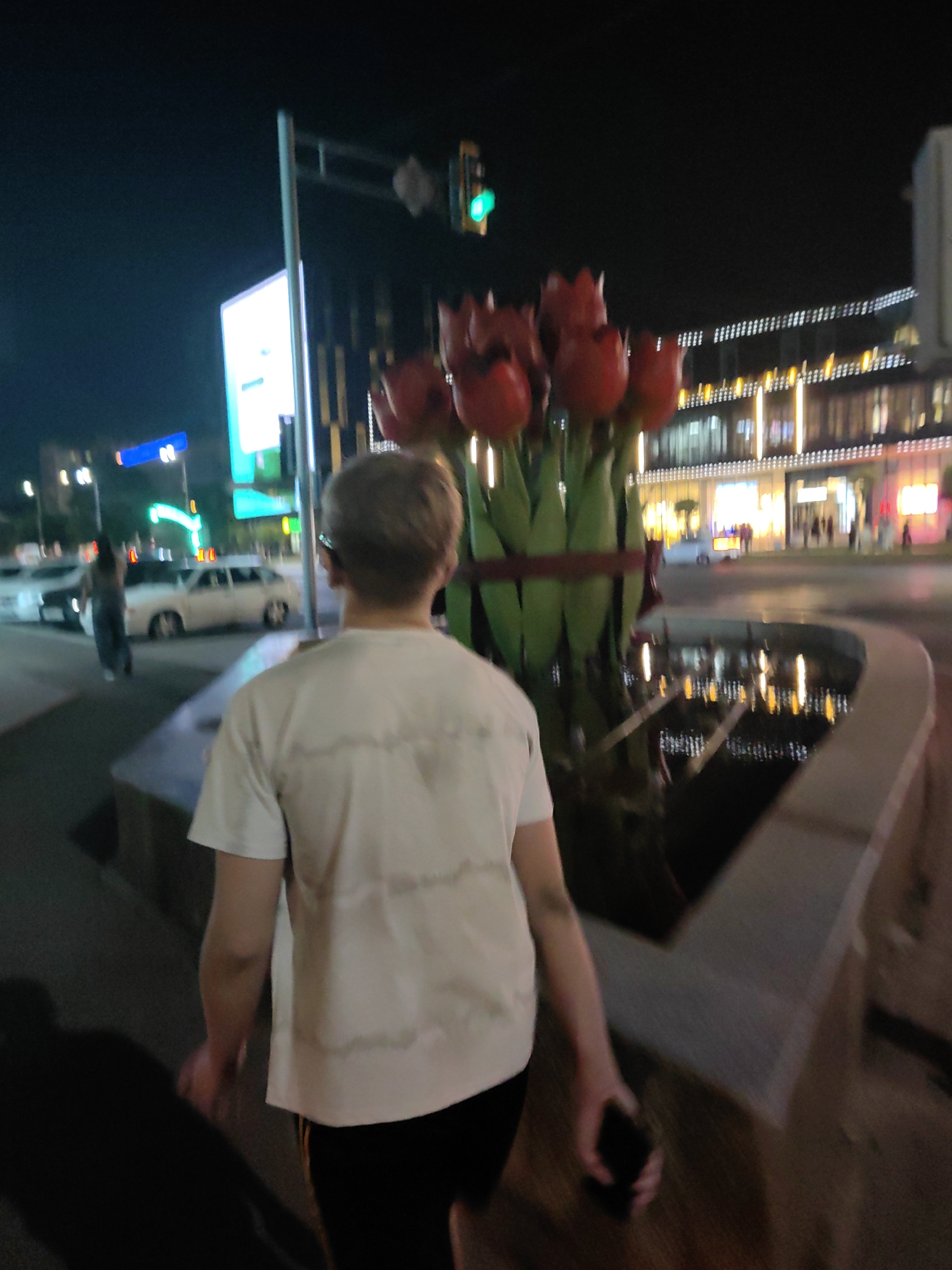 In Shymkent, Kazakhstan, at night, a person with blond hair walks away from the camera on a sidewalk. The person is wearing a white t-shirt and black shorts, and is holding a dark-colored smartphone in their right hand. Behind the person, a large artificial bouquet of red tulips stands in a fountain. In the background, cars and a woman in a gray dress are visible on the street. Further back, a brightly lit modern building with numerous windows stands out against the night sky. A traffic light with a green signal is also visible. The scene is characterized by artificial lighting from the building and streetlights, reflecting on the wet surface of the fountain.