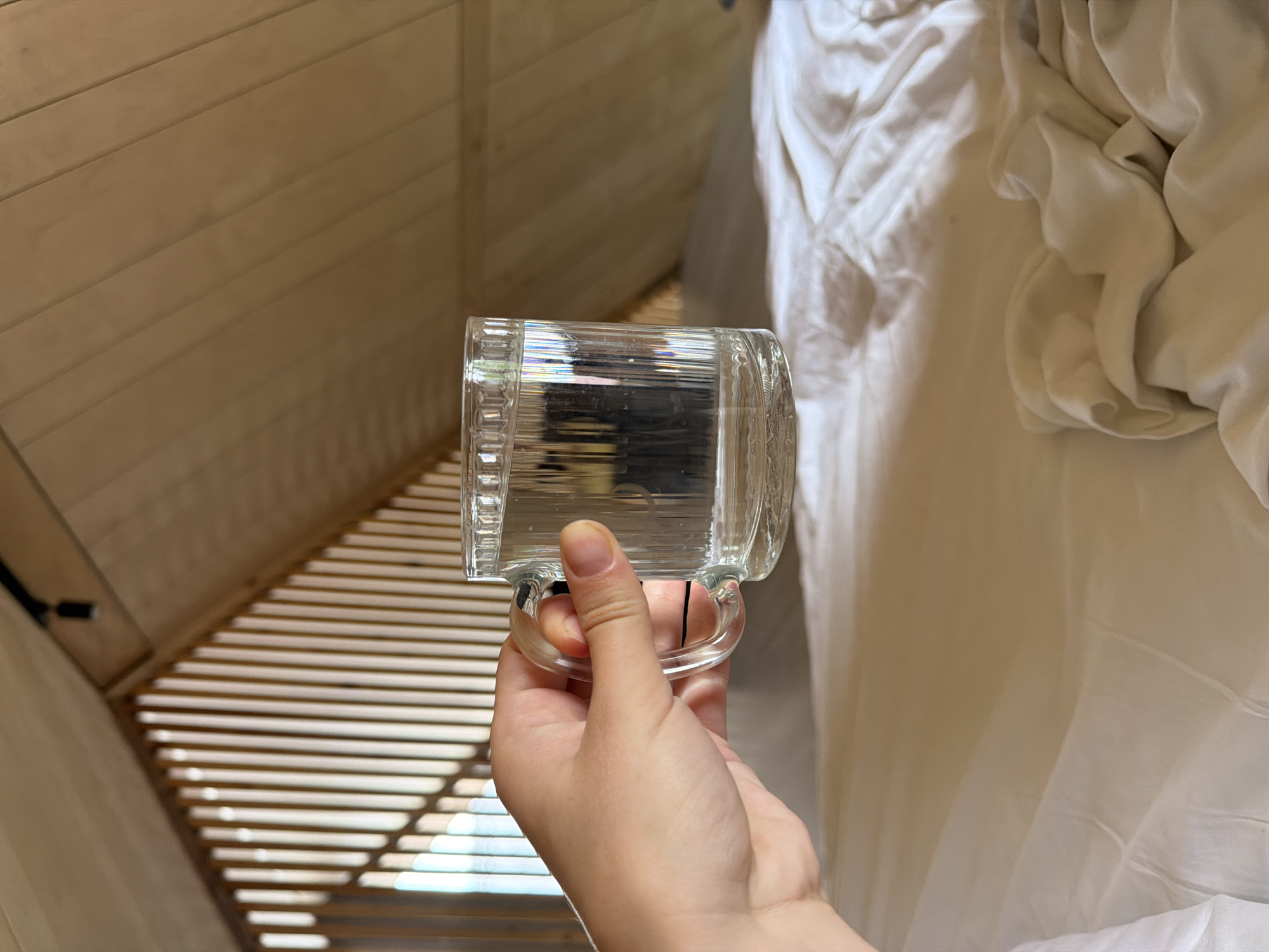 A hand holds a clear glass mug filled with clear liquid. The mug has vertical ridges and a curved handle. The background features a sloped wooden ceiling with visible planks and a black track light. To the left, a slatted wooden structure casts shadows on the ceiling. Below this, a white, rumpled bedsheet is visible. The scene is indoors and does not provide any information about the city of Семилуки, Russia.