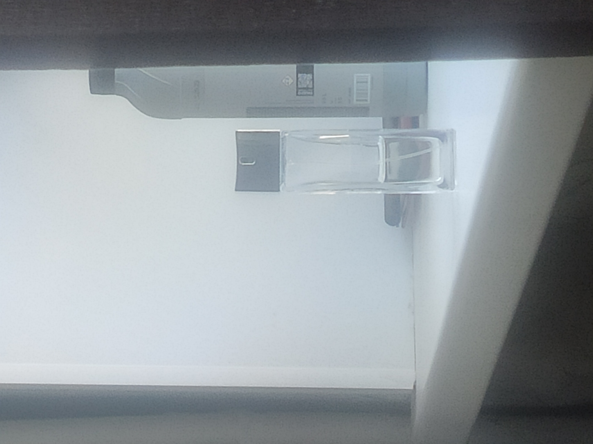 The image shows the interior of a drawer, likely a dresser or vanity drawer. A bottle of liquid, possibly cleaning solution, and a rectangular glass perfume bottle with a gray cap and atomizer are resting on the drawer's white surface. The edge of the drawer is visible, and the image is slightly rotated. The location is not discernible from the image.