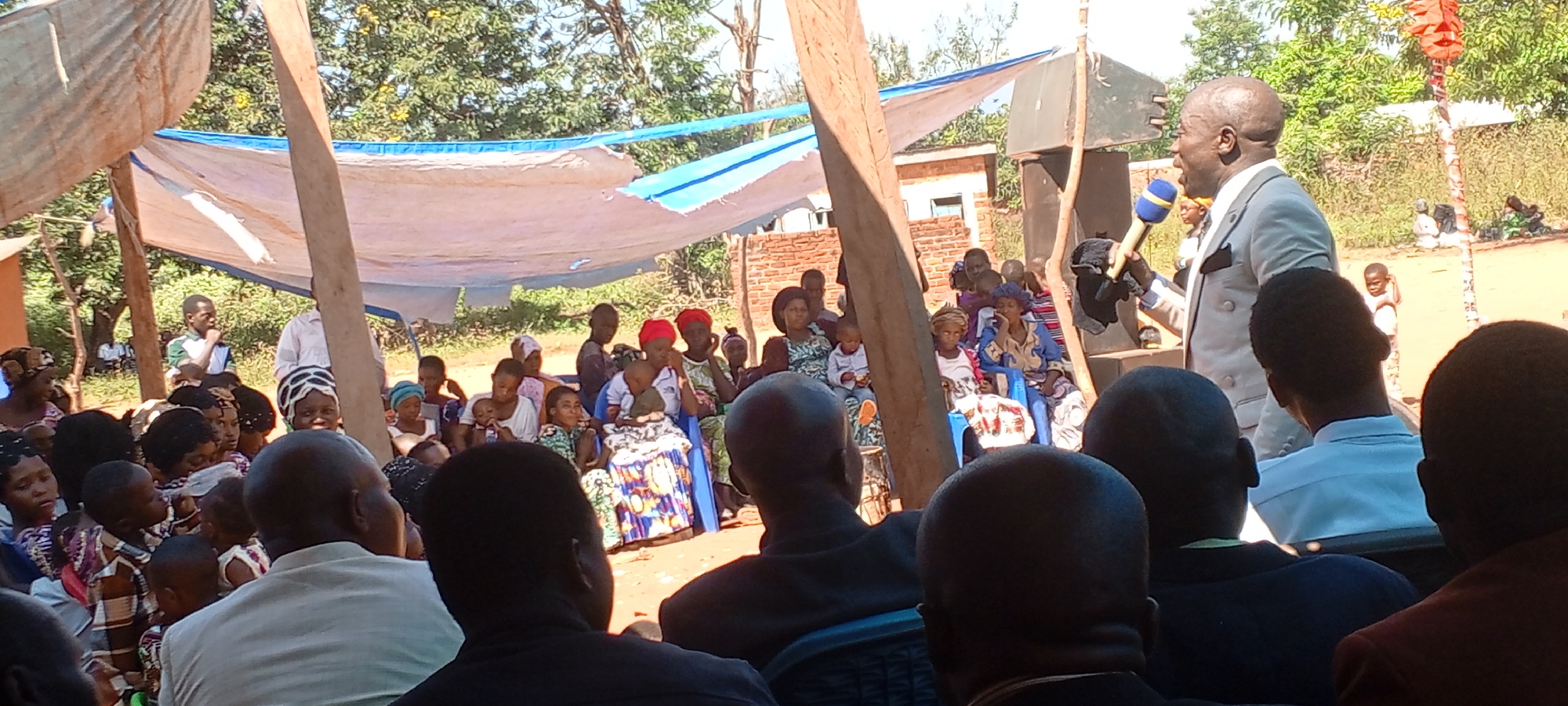 The image depicts a gathering of people in an outdoor setting, likely in a rural area, based on the presence of trees, a dirt ground, and simple structures. A temporary shelter, created with wooden poles and white cloth, provides shade. The location is identified as Kalemie, Democratic Republic of the Congo.
The focus is on a man in a light-colored suit speaking into a microphone. He appears to be the central figure addressing the group. People of various ages and genders are seated in rows facing the speaker. Some are seated on plastic chairs, and they seem to be listening to the man.
In the background, a building with a brick wall can be observed. Overall, the scene conveys a sense of community.