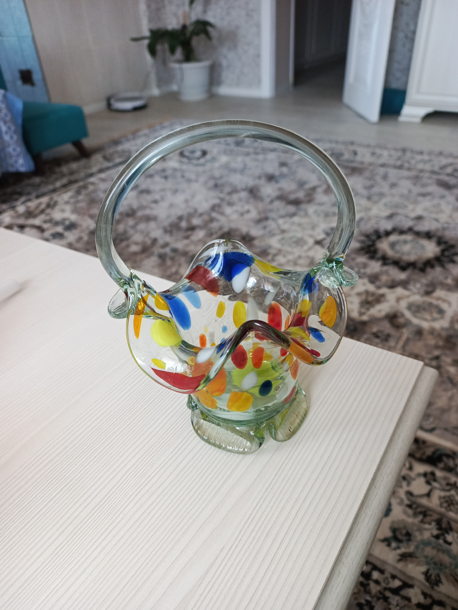 A colorful, clear glass basket sits on a white-colored, textured surface. The basket is decorated with multi-colored dots and features a circular handle. The background is blurry but appears to include a carpet, a chair, a houseplant, and a doorway. There are no visible people. Based on the prompt, this scene is located in Chaikovsky, Russia.