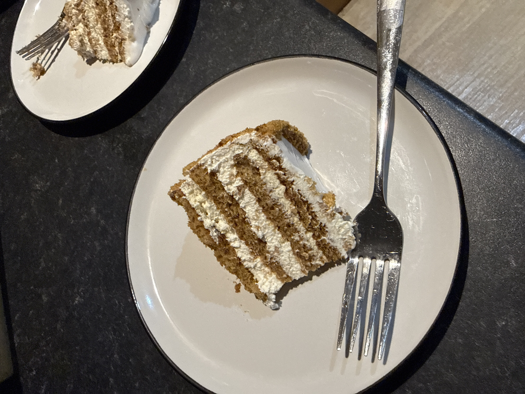 Two slices of layered cake, likely a sponge cake with cream filling, are presented on white plates. The cake on the left is a large triangular slice, featuring distinct brown cake layers alternating with thick white cream. A silver fork rests on the plate, with its tines piercing the edge of the cake slice. The plate itself is white with a thin dark rim. The second plate, partially visible in the upper right quadrant, holds a smaller portion of a similar cake, with a fork also present. Both plates sit on a dark, speckled countertop, possibly granite or a similar material. The lighting is dim, creating shadows and highlighting the textures of the cake and countertop. The scene appears to be an indoor setting, likely a kitchen or dining area, and there are no people visible. The presence of food and tableware suggests a meal or snack is being consumed. The specific location in Great Yarmouth, United Kingdom, cannot be determined from the visual information provided in the image.