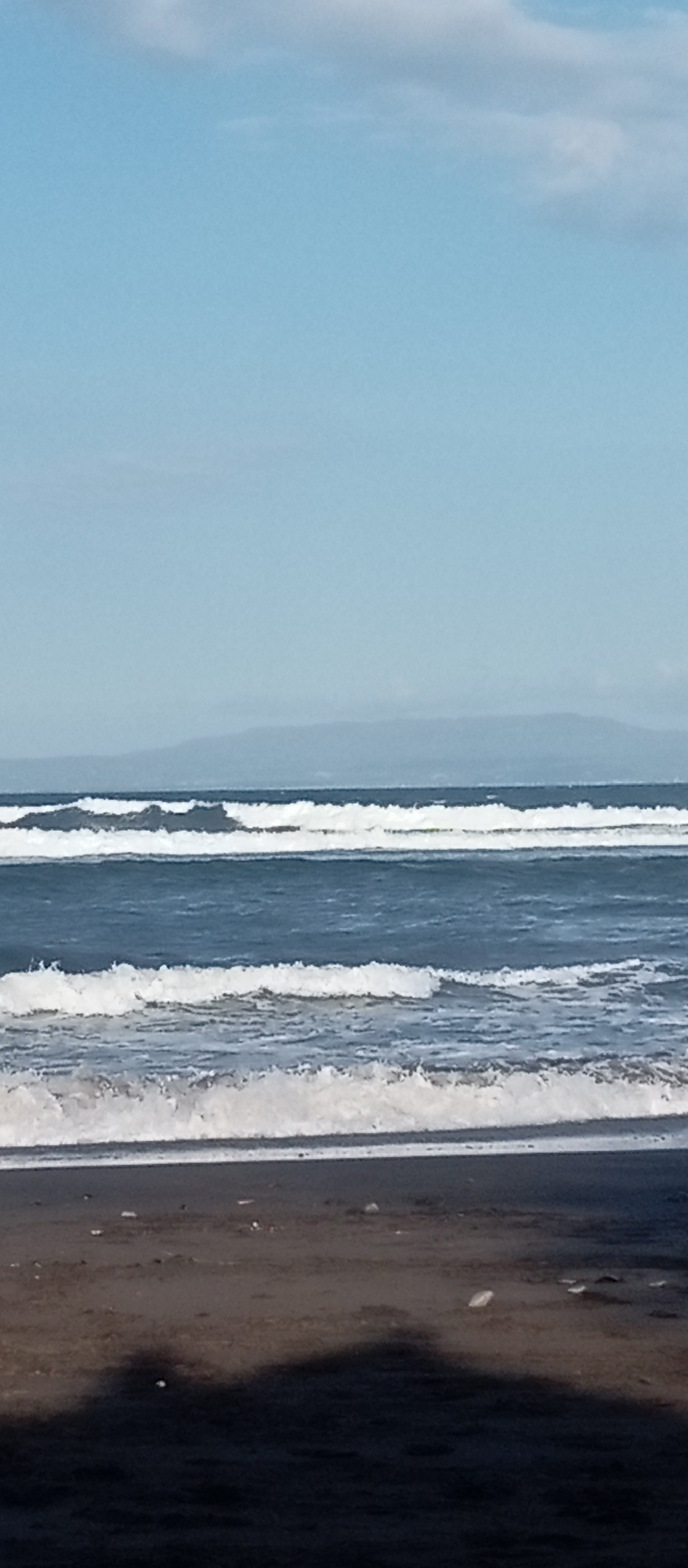 The image depicts a serene beach scene with the ocean, sand, and sky. In the foreground, dark, wet sand stretches across the bottom of the frame, with some scattered debris. The middle ground is dominated by the ocean, with several rows of white-capped waves rolling towards the shore. The water itself is a deep blue. In the background, a hazy, distant landmass is visible, likely a coastline or mountains, under a clear, pale blue sky with a few wispy clouds. The scene appears to be a natural coastal environment. The image does not contain any people or specific landmarks that would identify it as being in Blahbatuh, Indonesia.