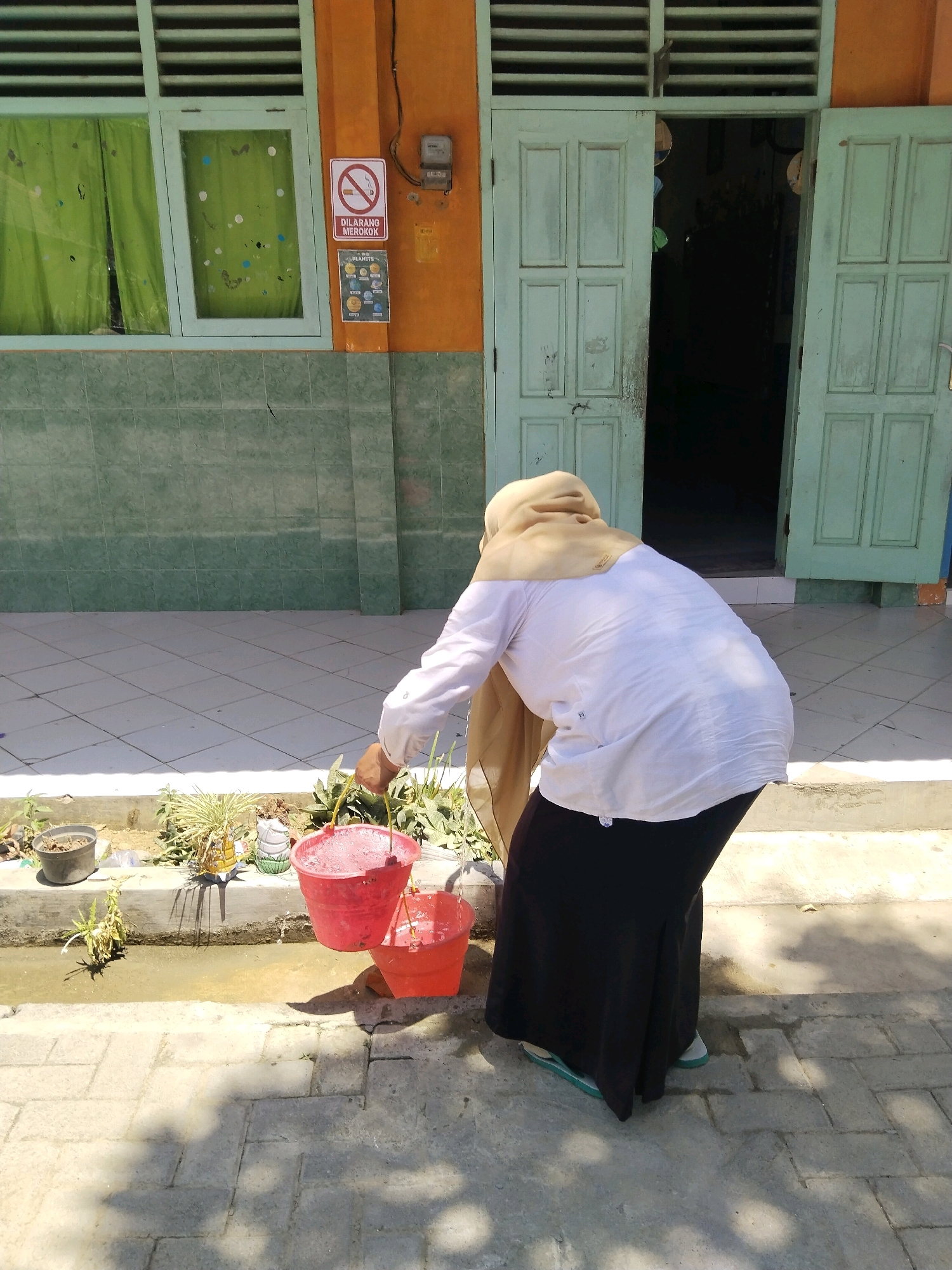 A person wearing a light brown hijab, a white long-sleeved shirt, and a black skirt is standing in front of a building with orange and green walls. The person is bent over and holding a red bucket in their right hand and an orange bucket in their left hand. The buckets appear to be filled with water. The building has a light blue double door that is partially open. To the left of the door, there is a window with green curtains. A sign that says "DILARANG MEROKOK" (No Smoking) is posted on the wall to the right of the window. The ground in front of the building is paved with light-colored tiles. There are some plants in pots to the left of the person. The scene appears to be outdoors in a sunny location.