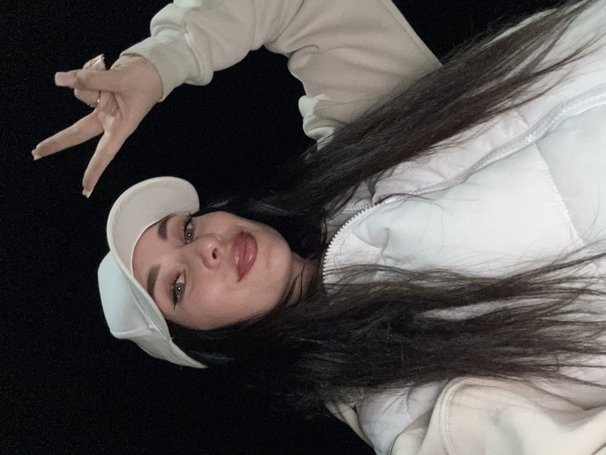This close-up, angled selfie captures a young woman in Chaikovsky, Russia, against a solid black background, suggesting nighttime or a dark indoor setting. Her head is tilted, and she gazes directly at the camera with a neutral to slightly playful expression. She has long, dark hair flowing around her shoulders and face. Her facial features include well-defined eyebrows, noticeable eyeliner creating a winged effect, and light-colored eyes. Her lips are slightly parted, highlighted with a reddish-pink gloss.
She wears a light-colored (off-white or cream) hooded top, layered beneath a matching quilted or puffy vest/jacket, which has a visible zipper. A light-colored baseball cap is worn on her head, positioned at an angle. Her right hand is raised towards the upper left of the frame, forming a peace sign with her index and middle fingers. Her fingernails are long and appear to have light-colored polish. No text is visible in the image.