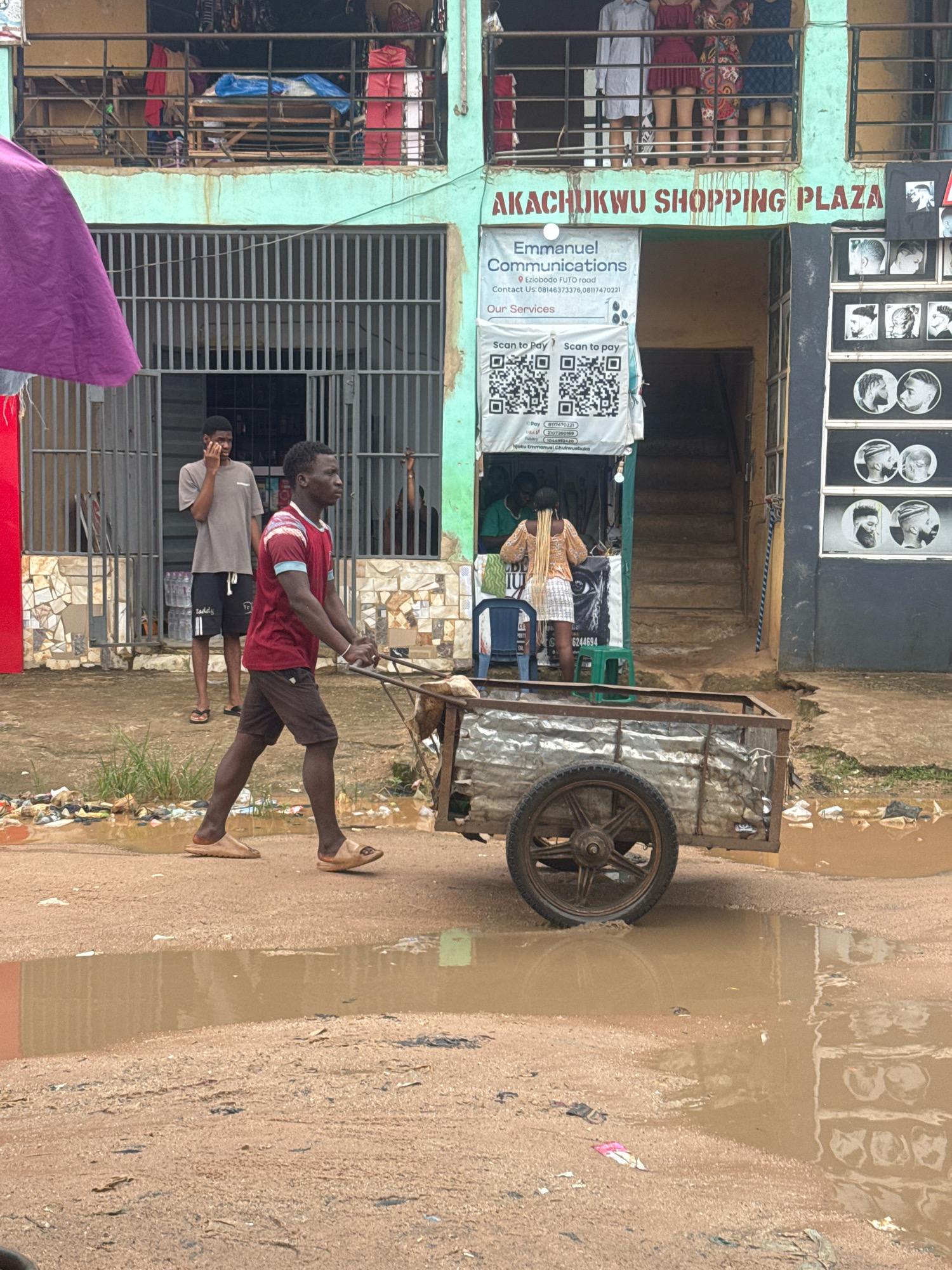 In Ihiagwa, Nigeria, a man is pushing a cart filled with items. The cart is on a dirt road with puddles of muddy water, indicating recent rain. In the background, there is a building with a sign that reads "AKACHUKWU SHOPPING PLAZA." The building appears to be a market or commercial center, with several shops and people visible. One shop displays pictures of hairstyles, likely a barbershop. The overall scene suggests a bustling urban environment with everyday activities taking place.