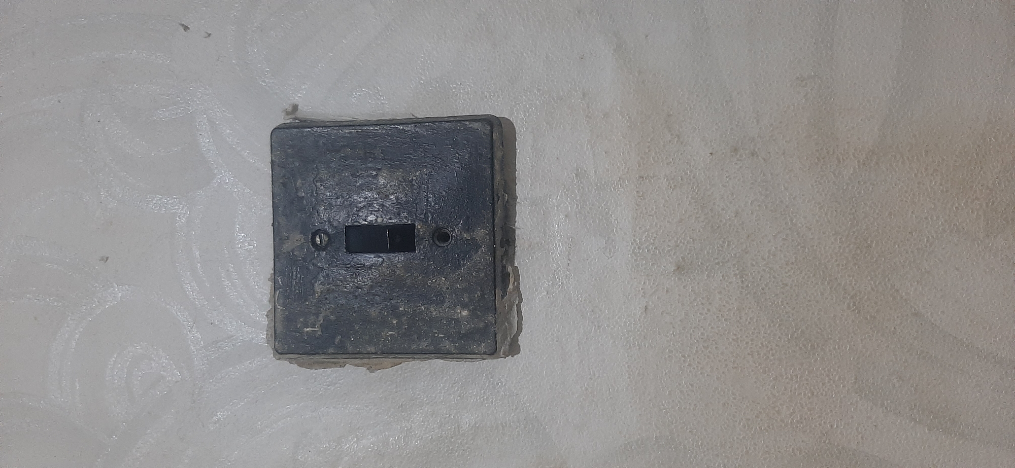 A close-up, eye-level shot shows a weathered, black, square electrical light switch mounted on a pale, textured wall in Yessentukskaya, Russia. The switch has a rectangular button in the center, with a visible line dividing it into two smaller sections, indicating it likely controls two separate circuits or lights. The switch plate is visibly dirty and worn, with chipped edges and a rough texture, suggesting age and considerable use. Two circular indentations, likely for screws, are visible on either side of the switch button, further highlighting its utilitarian design. The surrounding wall is a light, off-white color with a faint, swirling pattern etched into its surface, possibly from past repairs or decoration. The texture of the wall is slightly bumpy and uneven, with small, circular indentations scattered across it, creating a mottled appearance. The lighting in the image is even and appears to be artificial, with no strong shadows or highlights, suggesting an indoor setting, likely in a building. The scene is simple and functional, focusing entirely on the details of the aged electrical switch and the wall it is mounted on, devoid of any other objects or human presence. There is no visible text in the image.