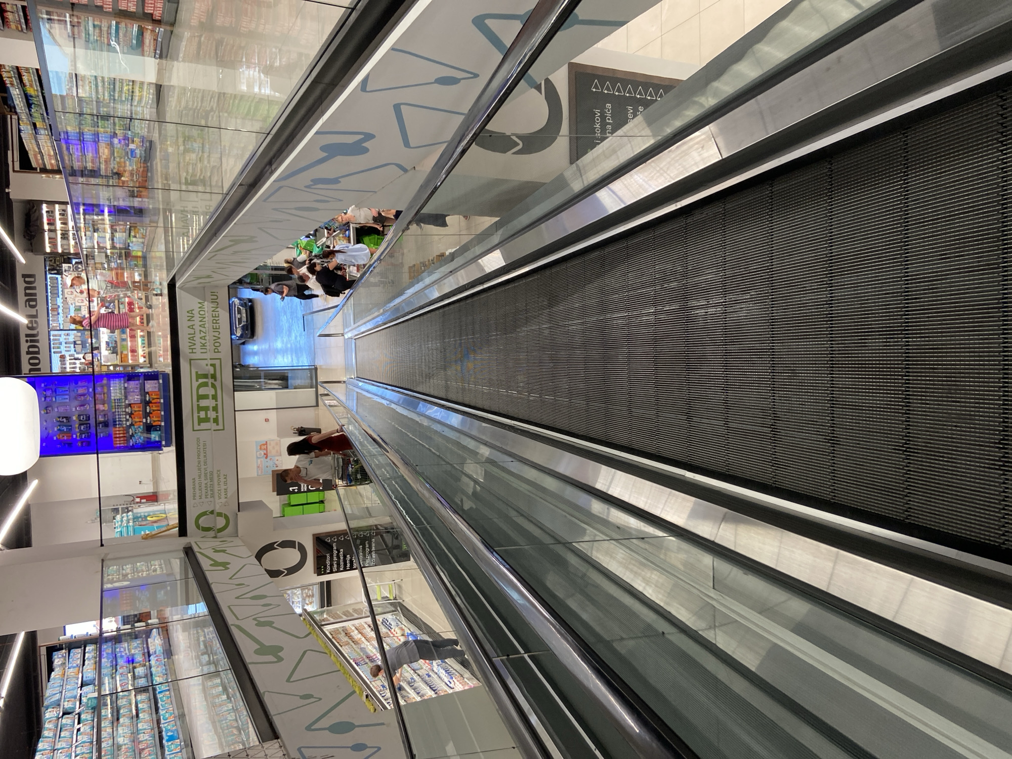 The image depicts a moving walkway inside a shopping center or supermarket, likely in Budva, Montenegro, given the presence of Montenegrin text on signage. The escalator ascends from the foreground towards the background. Shoppers are visible at the top of the moving walkway. The shopping center features reflective surfaces, displaying a mirrored view of shelves stocked with groceries and consumer goods. The walkway's sides are enclosed by glass panels. Signage with product categories and commercial messages is displayed along the sides of the escalator.