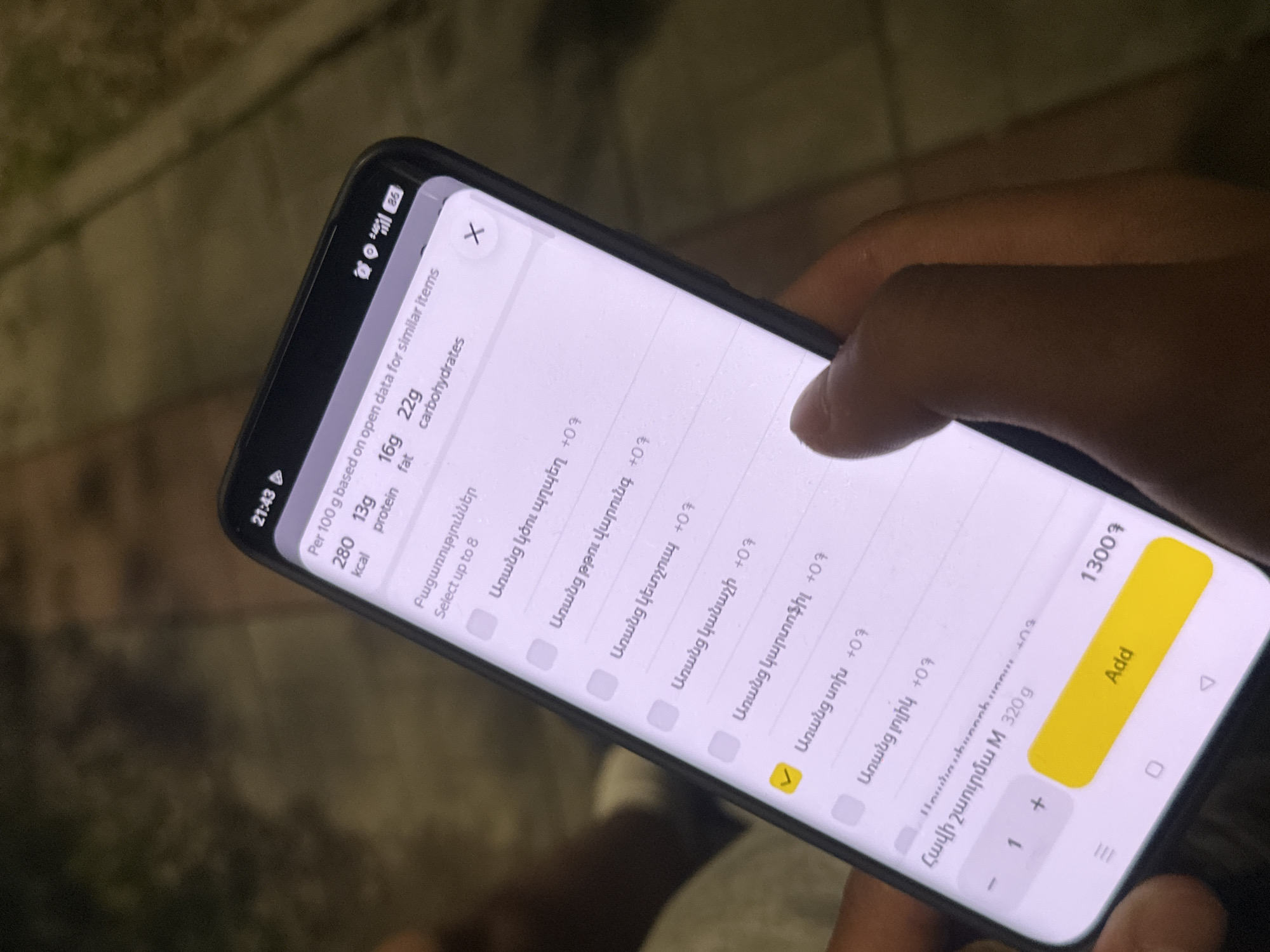 A person is using a smartphone, possibly for ordering food online. The screen displays a food ordering interface with nutritional information and options to customize the order. The order shows the food items in Armenian. The time shown on the phone screen is 21:43. The location is possibly Yerevan, Armenia, based on the Armenian language in the app. The user is touching the screen, presumably to confirm or customize the order. The "Add" button is highlighted in yellow, and a price of "1300?" is visible.