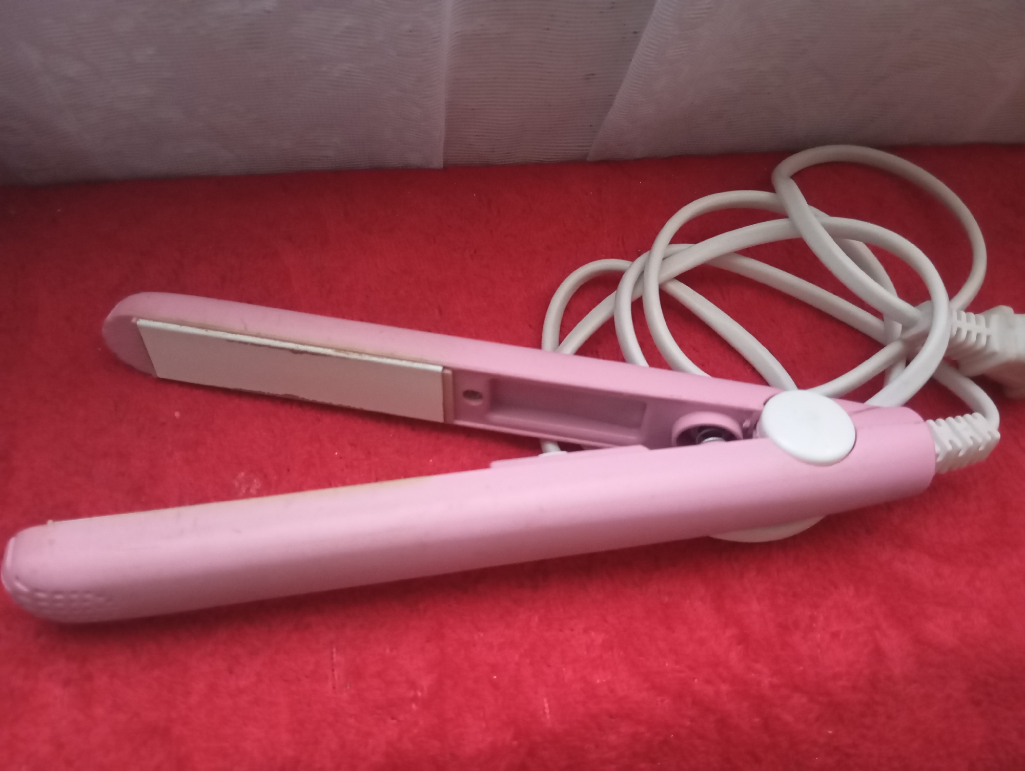 A pink hair straightener with a white cord and plug rests on a red textured surface. The hair straightener is partially open, revealing its heated plates. The background consists of a light-colored, ribbed material. The scene appears to be indoors, with no discernible indication of a specific location in Кош-Дөбө, Kyrgyzstan.