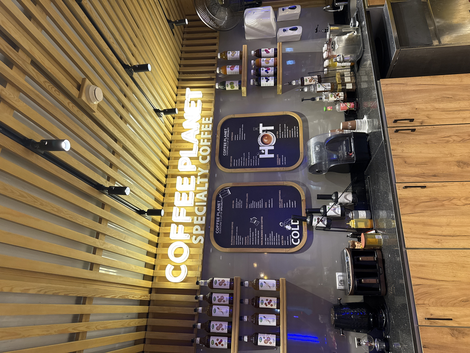 A coffee shop counter is captured in a medium shot, showcasing a variety of products and equipment. The scene is set in a brightly lit establishment with a wooden slatted ceiling and modern lighting fixtures.
Dominating the background is a prominent illuminated sign that reads "COFFEE PLANET SPECIALTY COFFEE". To the left, shelves are lined with multiple bottles of flavored syrups, each with a white label featuring illustrations.
In the center, a menu board with a black frame lists various coffee and drink options, categorized under "COFFEE PLANET" and "EVERYONE'S CUP OF SPECIALTY". The menu details include "BLENDED ICED COFFEE", "FRAPPE", "NON BLENDED ICED COFFEE", "HOT COFFEE", "CHOCOLATE", "HOT TEA", and "FRUIT SMOOTHIES". A prominent graphic of a steaming cup of coffee is featured within the menu.
To the right of the menu board, more shelves hold an assortment of bottled beverages with colorful labels, some featuring images of fruits.
The counter itself displays a coffee machine, a kettle, and various other coffee-making accessories, including syrups, dispensers, and what appear to be cups. A large industrial fan is mounted on the wall behind the counter to the right. The lower portion of the image reveals wooden cabinet doors with black handles, part of the shop's interior design. The overall impression is that of a well-stocked and modern coffee shop, likely in a commercial area given the professional presentation.