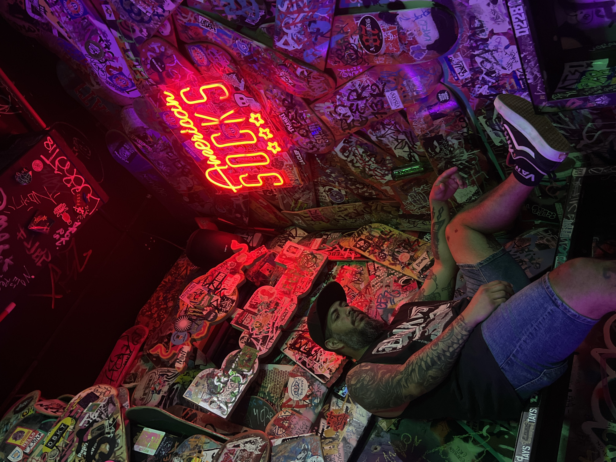 An eye-level, tilted photograph captures an immersive, alternative indoor space, likely a bar or creative venue, in Barcelona, Spain. The ceiling and walls are entirely adorned with a dense collage of skateboard decks, heavily covered in an array of colorful stickers and graffiti, creating a vibrant, textured backdrop. The scene is bathed in dim, atmospheric lighting, primarily emanating from red and purple neon signs and ambient illumination.
A man is depicted lying relaxed amidst this unique environment, his head resting on the skateboard-covered surface, and his legs casually bent, with one foot visible. He is wearing a black t-shirt, denim shorts, a black baseball cap, and black Vans sneakers. Tattoos are prominently visible on his arms and legs. His posture conveys a sense of calm and leisure within the energetic setting.
A bright yellow-orange neon sign spelling "American SOCKS" with three stars serves as a central focal point. Numerous other pieces of text, primarily from stickers and graffiti, are visible across the skateboard surfaces, including "VANS" (on the shoe), "HELLO," "TAYS," "DSKT," "MINY," "TRANKI," "VISADO," "Red Bull," "LACLEF," and various other tags and phrases such as "MELODICA" and "CHAT," contributing to the rich, layered aesthetic. The overall ambiance is dark, cool, and distinctly urban.