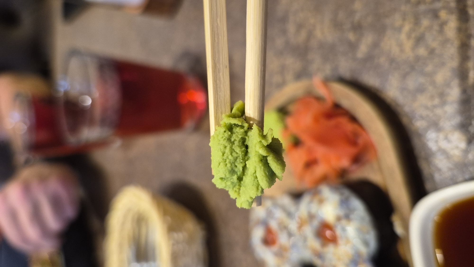 A close-up, eye-level shot shows a dollop of vibrant green wasabi being held by two wooden chopsticks. The wasabi is textured and appears freshly prepared. In the background, out of focus, are elements of a meal. To the left, a basket-like dish with a light-colored interior is visible. Further back, two glasses filled with a reddish liquid, possibly a drink, are present. On a wooden platter, pink pickled ginger is arranged next to what seem to be sushi rolls. A small white dish containing a dark liquid, likely soy sauce, is in the lower left corner of the frame. The setting suggests a restaurant or dining environment. No specific location in Daugavpils, Latvia can be identified from the image.