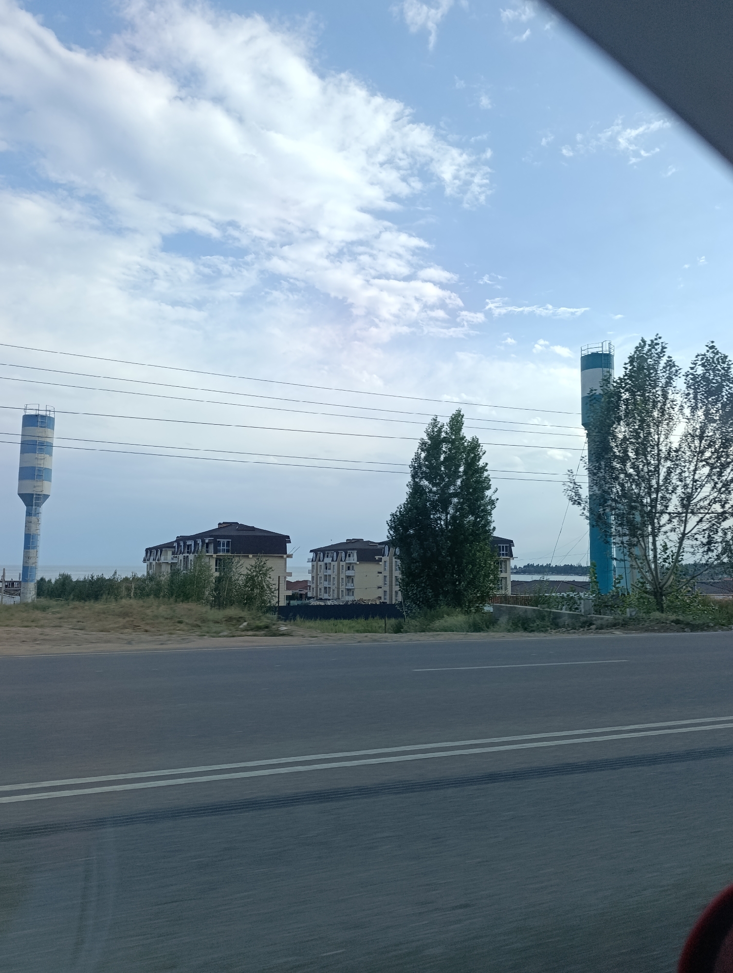 The image depicts a daytime exterior scene. In the foreground, a grey asphalt road with white double lines runs horizontally across the frame, showing tire tracks. Beyond the road, a grassy embankment with some sparse vegetation leads to a row of modern apartment buildings with dark-pitched roofs. To the left of the buildings, a tall, cylindrical water tower with blue and white horizontal stripes stands against a hazy blue sky with scattered white clouds. Further to the right, two tall trees with lush green foliage are visible, with another similar water tower partially obscured behind the rightmost tree. In the background, a body of water, possibly a lake or sea, can be seen beyond the buildings, with a distant hazy shoreline. Power lines are strung across the middle ground, running horizontally. There are no people visible in the image. The scene appears to be on the outskirts of a settlement.