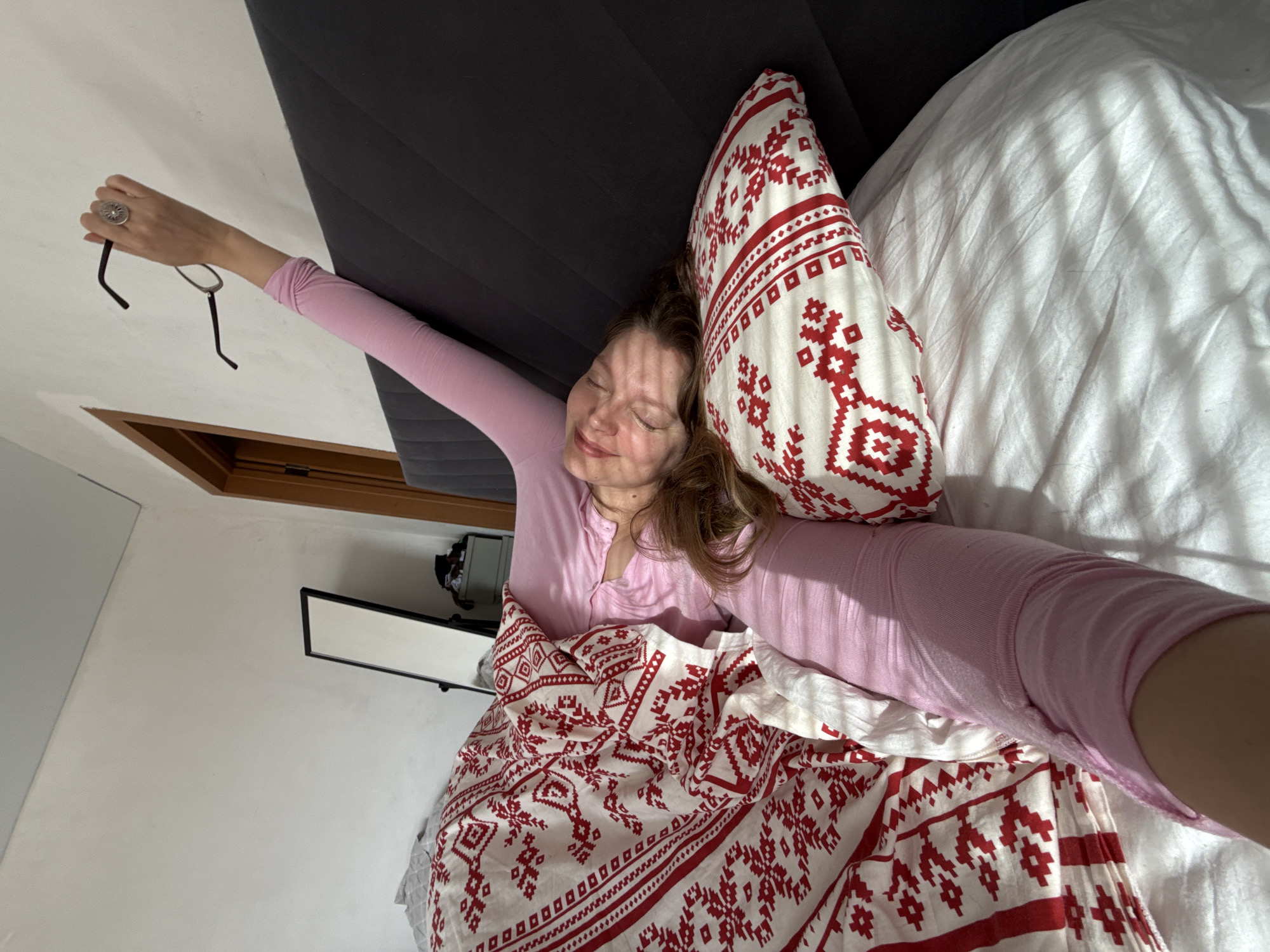 A woman is lying in bed under a red and white patterned duvet. She wears a pink long-sleeved shirt. Her right arm is raised and she holds a pair of glasses. A pillow with a matching red and white pattern supports her head. A second white pillow is adjacent. The bed has a dark grey headboard. A mirror is mounted on the white wall. The location is not evident in the image. There is no information to suggest this is in Izhevsk, Russia.