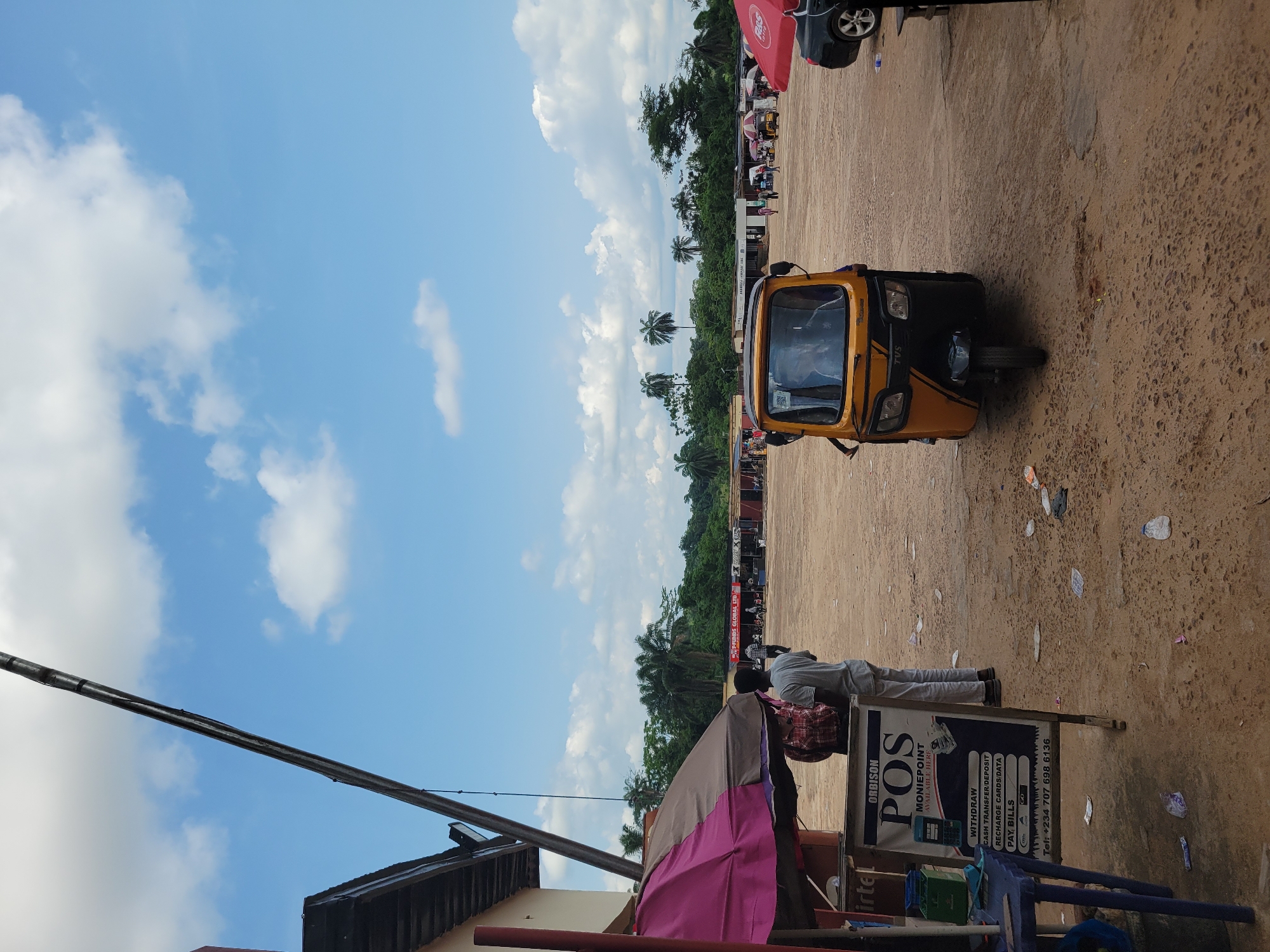 The image depicts an outdoor scene in Egbagu, Nigeria, characterized by a dusty, open area with scattered debris on the ground. In the foreground, a yellow and black auto-rickshaw (a TVS model) is parked. To the left, a sign for "ORBISON POS MONIEPOINT" is displayed, listing services like withdrawals, cash transfers, deposits, recharge cards, data, and bill payments, along with a phone number. Behind the sign, an umbrella with pink and brown fabric is partially visible. A person in a grey shirt and white trousers stands near the sign. Further in the background, a line of small shops or stalls with corrugated metal roofs are visible, some displaying signage such as "FRIENDS GLOBAL LTD" and "BAHWEBA COMPANY LTD". Several people are seen walking between the stalls. A black car is parked on the right side of the image. The sky above is a clear blue with scattered white clouds, and palm trees and dense green foliage form a backdrop behind the stalls. A large metal pole, possibly part of a utility structure, extends diagonally across the upper left portion of the image. The overall impression is of a busy, open-air market or commercial area in a rural or semi-urban setting.