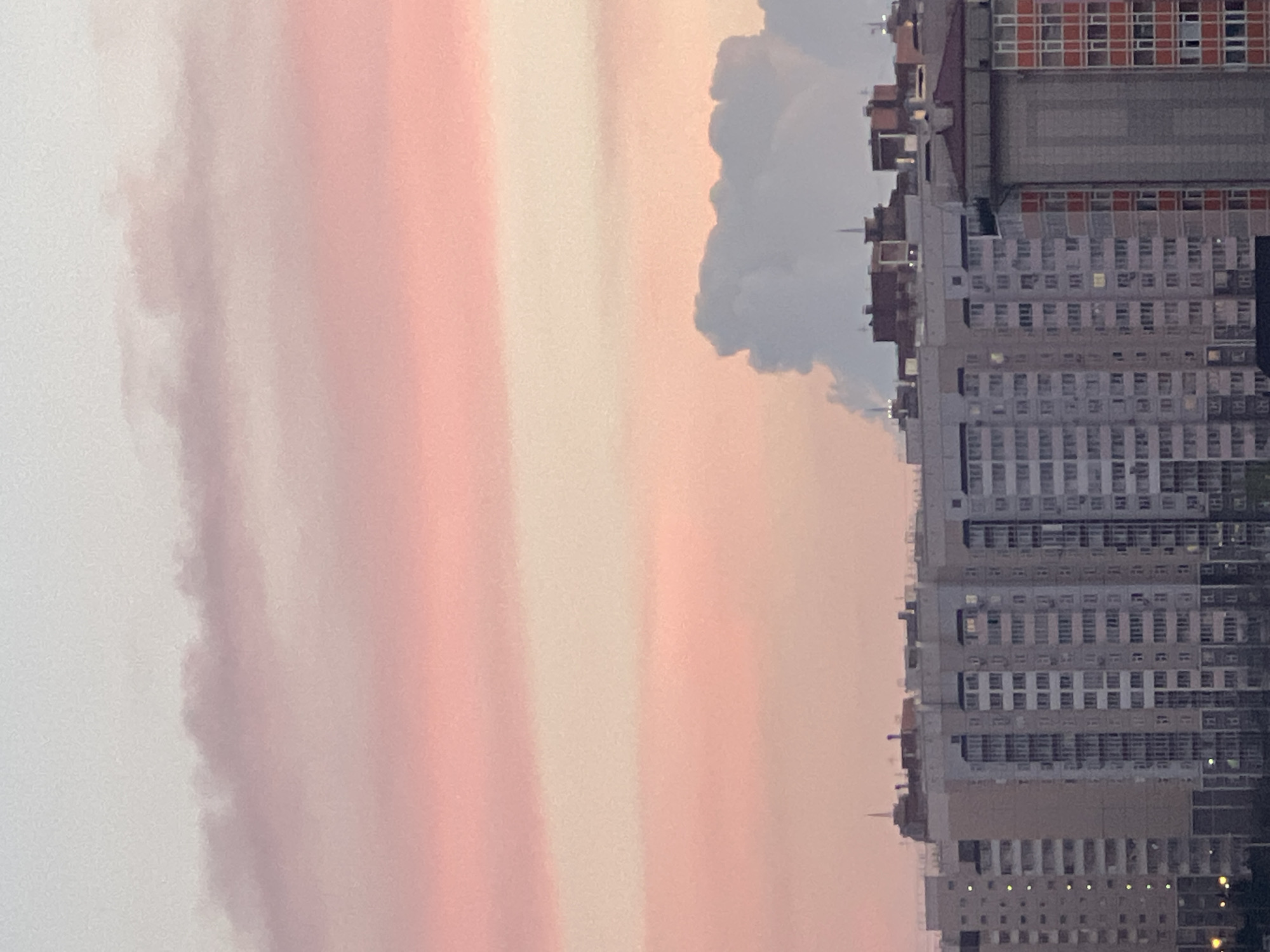 The image shows a high-angle view of a building complex in what appears to be Krasnoyarsk, Russia, based on the architecture. The buildings are mostly high-rise residential structures with a regular pattern of windows. The sky is filled with streaks of pink and orange light, indicating a sunset or sunrise. There are gray clouds near the buildings. No people are visible in the image.