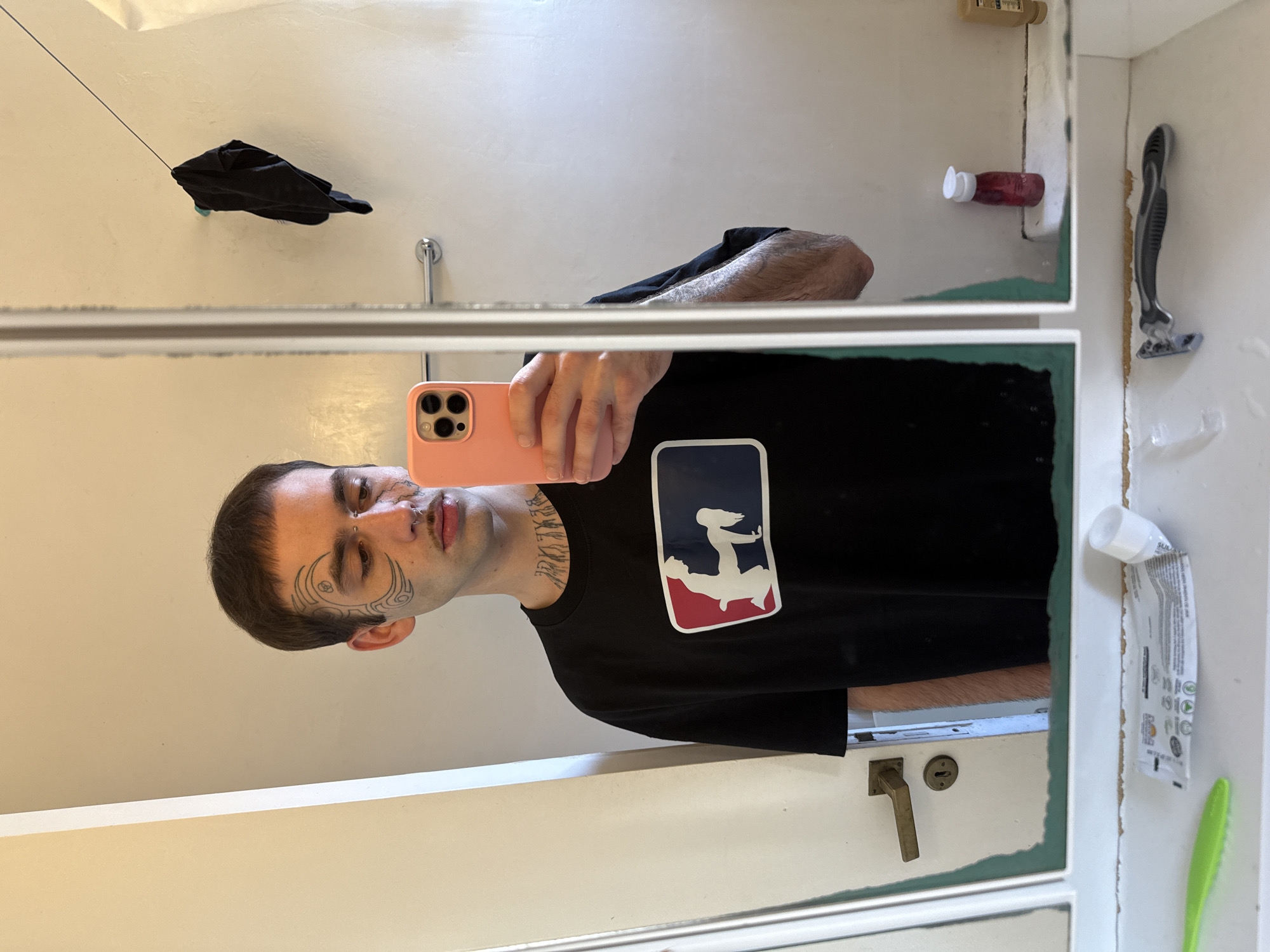 The image is a mirror selfie, captured in a bathroom and rotated 90 degrees clockwise. It features a young adult male with short, dark hair and prominent facial tattoos, including swirling patterns around his left eye. He has a nose ring, possibly other piercings, and a mustache with stubble. His expression is neutral.
He is holding a light peach-colored smartphone, equipped with multiple camera lenses, to take the photo in the mirror. He is wearing a black t-shirt that displays a rectangular logo with red, white, and blue sections. Inside the logo, white silhouettes depict two figures in a dynamic pose, reminiscent of the NBA logo. Visible text on his neck and collarbone area, likely a tattoo, reads "BEXLEY LA".
The bathroom setting is reflected around him. Visible items include a black garment hanging from a thin string, a small red bottle with a white cap, and a grey and black razor. A partially squeezed white tube, possibly toothpaste, is visible on the right, next to a green brush. The mirror appears to be part of a cabinet, characterized by its edges and green trim, which shows some signs of wear and chipping. Below the mirror's reflection, a brass door handle and keyhole are present. The white tube has small, partially legible text, including "SULI" at the top.