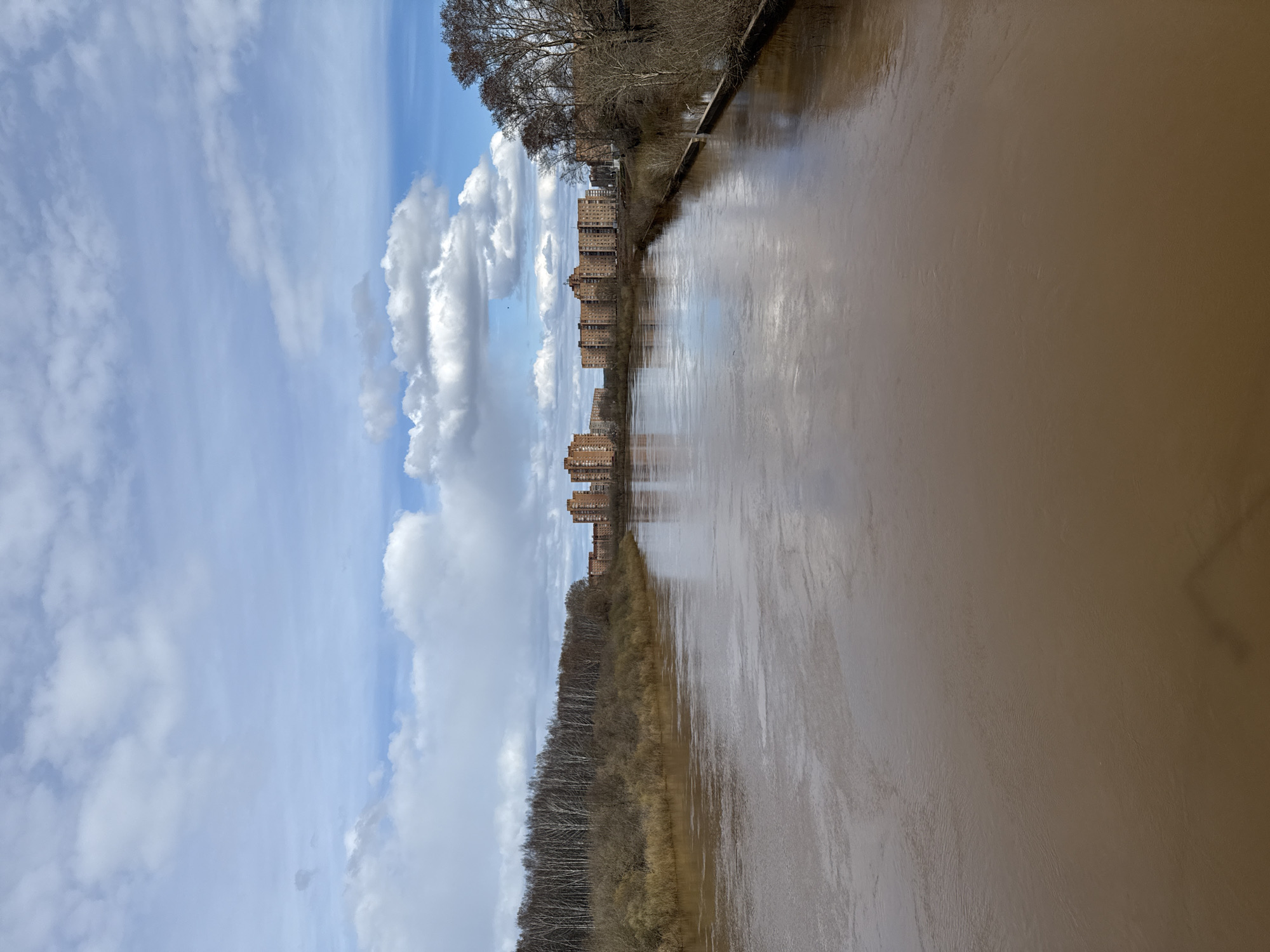The image captures a panoramic view of a wide river flowing through a landscape in Glazov, Russia, under a dynamic, partially cloudy sky.
The river, characterized by its distinct muddy reddish-brown water, occupies the foreground and a significant portion of the right side of the frame. Its surface is calm, reflecting the bright blue sky, the fluffy white clouds, and the buildings lining the opposite bank.
On the left bank, a dense line of bare, deciduous trees, indicating late winter or early spring, forms a natural barrier. Beyond these trees, a long row of multi-story residential buildings with brick-like facades stretches into the distance, suggesting urban development along the river. Their reflections are visible in the water.
The sky, dominating the upper left, displays patches of clear blue interspersed with large, cumulus clouds and thinner, wispy clouds. The lighting suggests it is daytime, likely midday, with bright, diffused light.
The scene is devoid of visible human activity or discernible individuals. The overall impression is one of a serene, expansive landscape that juxtaposes natural riverine features with urban structures. No text is visible in the media file.