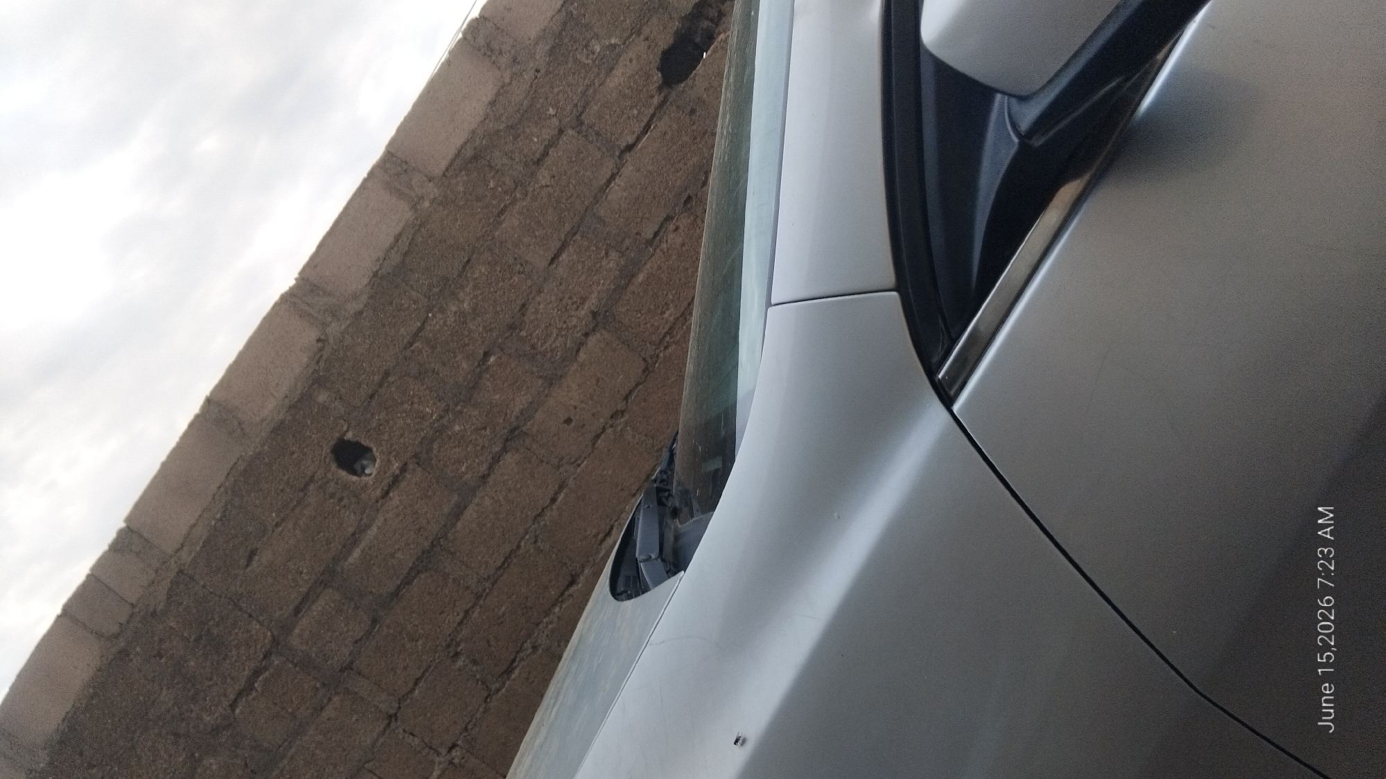 The image shows a close-up of the front of a silver car, with the driver's side hood and windshield wipers visible. The car is parked in front of a textured brick wall. The sky is overcast. The timestamp "June 15, 2026 7:23 AM" is visible in the bottom left corner of the image. There are no people or actions depicted in the image. The scene is static and appears to be an outdoor shot. The location is not specified as Howayi, Nigeria.