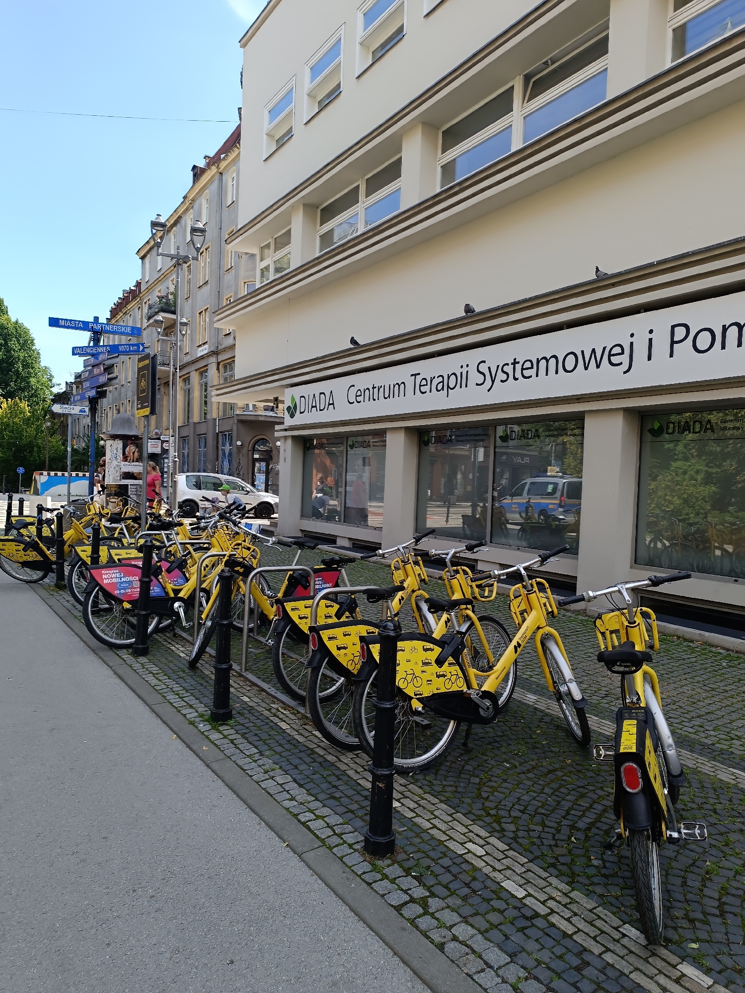 The image depicts a street scene in Gliwice, Poland, specifically in front of a building with a sign reading "DIADA Centrum Terapii Systemowej i Pomocy Psychologicznej." A row of bright yellow rental bicycles is parked along the sidewalk, secured by black metal posts. The bicycles have black seats, handlebars, and frames, with yellow accents on the fenders and frames. Some of the bicycles have informational stickers attached, including one with the text "Kongres Młodej MOBILNOŚCI."
Several street signs are visible in the background, including one pointing towards "MIASTA PARTNERSKIE" and another indicating "VALENCIENNES 1070 km." A few people are present in the background, including a woman in a pink shirt and a person wearing a green helmet. A white car and a police car are parked on the street. The sidewalk is paved with cobblestones, and the street itself is asphalt. The buildings in the background are typical of European city architecture, with multiple windows and light-colored facades. The sky is clear and blue, indicating a sunny day.