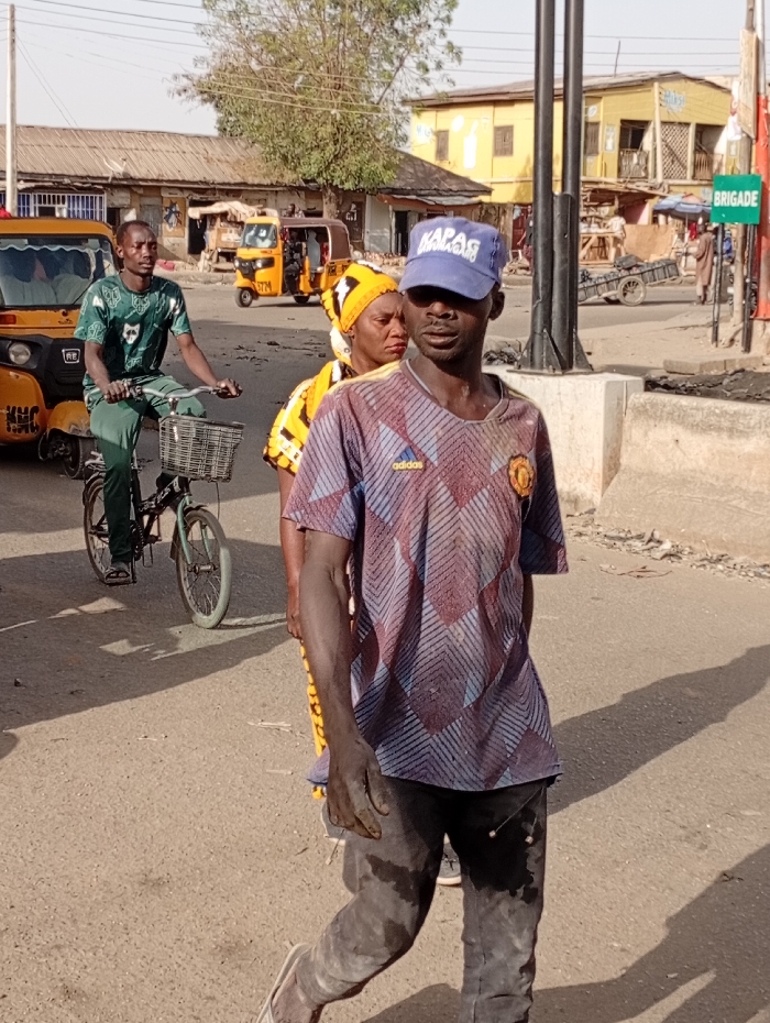 The image captures a vibrant street scene under bright daylight, likely in a town or city, featuring several individuals engaged in daily activities.
In the foreground, slightly to the right, a dark-skinned man is walking towards the left side of the frame. He is wearing a blue baseball cap with the word "KAPAS" embroidered in white letters on the front. He has on a short-sleeved t-shirt with a distinctive geometric pattern of maroon, blue, and white segments. A small white "adidas" logo is visible on the right chest area, and a circular crest, possibly a sports team emblem, is on the left chest. His trousers or jeans are dark and appear heavily soiled or dusty, particularly on the lower legs. His exposed right arm and hand also show signs of dirt or dust. He is wearing white flip-flops and looks slightly to his right, past the viewer.
Directly behind this man, a dark-skinned woman is partially visible. She is wearing a yellow headscarf with a patterned design and a yellow top or dress, also with a pattern. She is looking in a similar direction to the man in front of her.
On the left side of the frame, a dark-skinned man is riding a bicycle. He is wearing a green short-sleeved t-shirt with a lighter green or white pattern and matching green trousers. His bicycle is green and has a black basket attached to the handlebars. He is looking forward as he rides. A yellow three-wheeled vehicle, resembling an auto-rickshaw or tuk-tuk, is parked behind him, with "KHC" visible on its side.
The background reveals a bustling street environment. Further down the road, another yellow three-wheeled vehicle with "STM" and "S74" on its side is visible. A row of low-rise buildings with corrugated metal roofs lines the street, some painted in shades of yellow or light brown. The overall impression is that of an older, perhaps less developed, urban area.
To the right side of the image, a dark pole stands upright, and a green rectangular sign with the word "BRIGADE" in white capital letters is attached to another pole further down the street. A handcart or pushcart can also be seen in the distance near the buildings on the right. The road surface is asphalt, and prominent, elongated shadows are cast on the ground by the people and objects, indicating that the sun is high in the sky, suggesting it is mid-morning or afternoon. The weather appears clear and sunny.