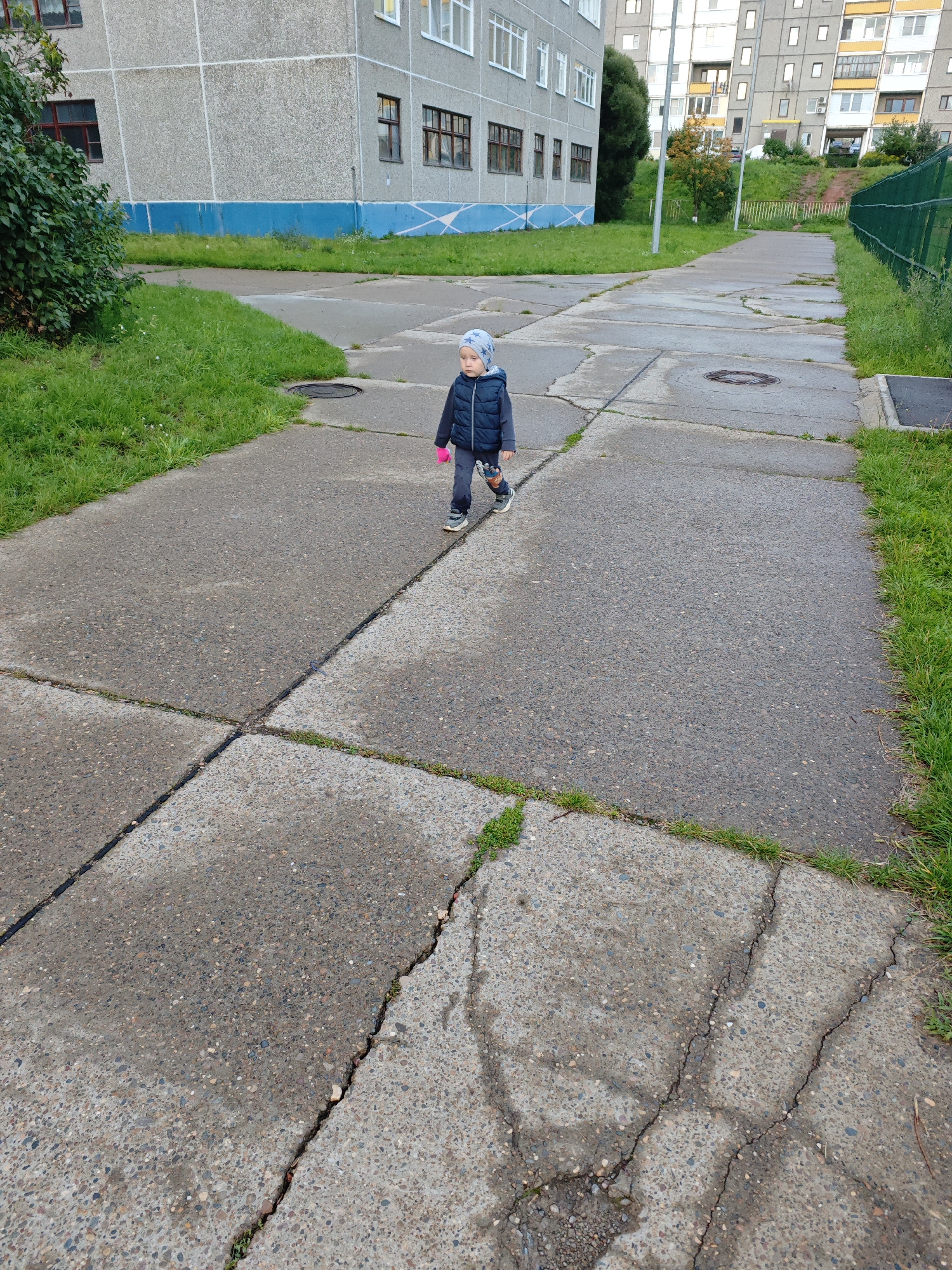 A young child, possibly a toddler, walks along a cracked concrete path with green grass growing between the slabs. The child is wearing a blue puffy vest, dark pants, a blue hat with stars, and pink gloves. They are carrying a toy in their right hand. In the background, a grey apartment building with blue trim is visible, along with other buildings and a green fence. The setting appears to be an urban environment in Markovskii, Russia, likely a residential area with shared pathways and green spaces. The ground is wet, suggesting recent rain.